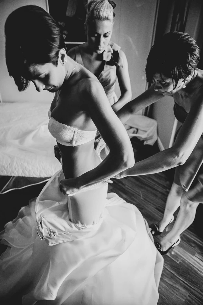 The bride being dressed by two women, standing by the bed.