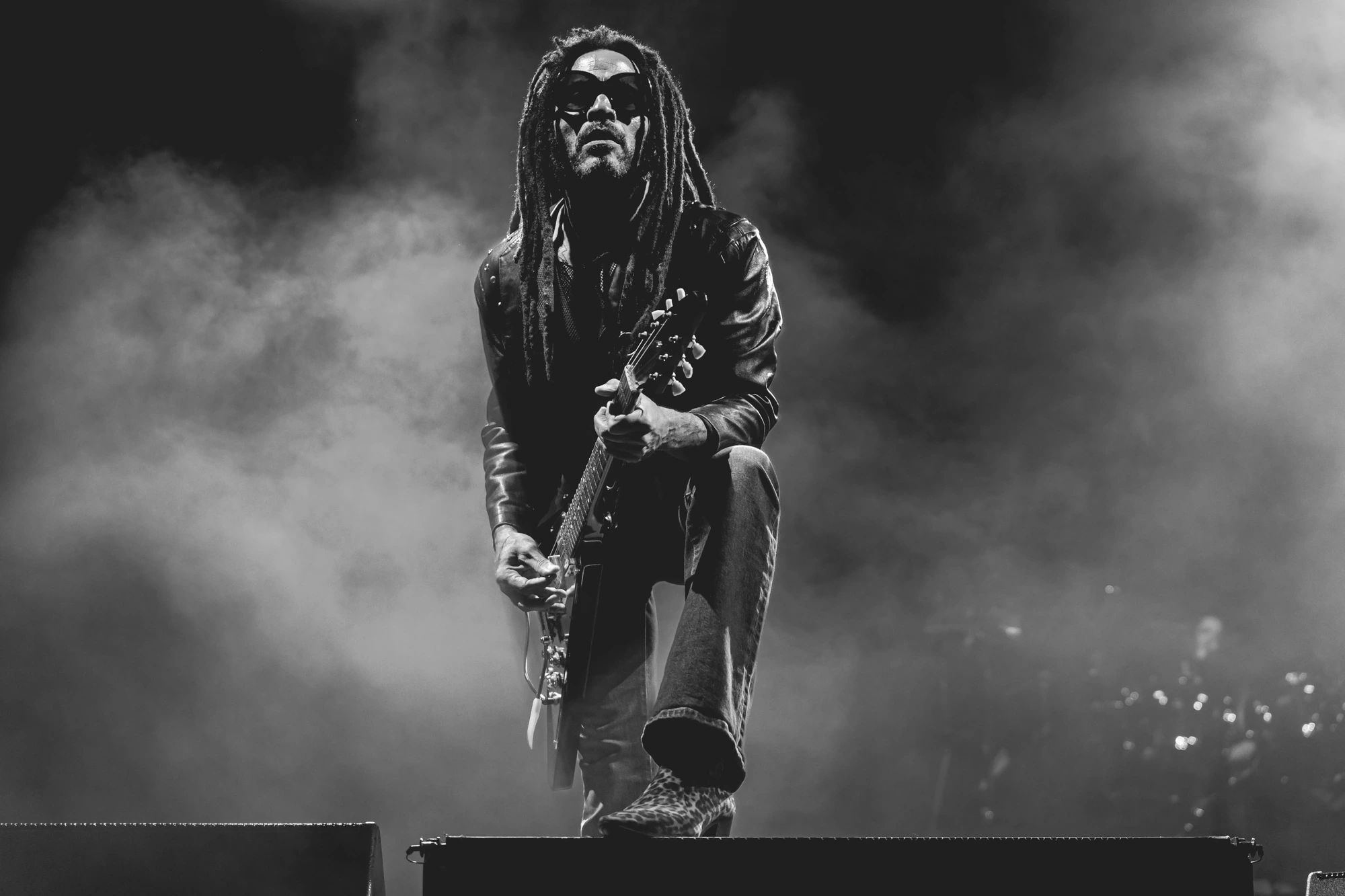 In black and white, a guitarist with dreadlocks wrapped in stage smoke.