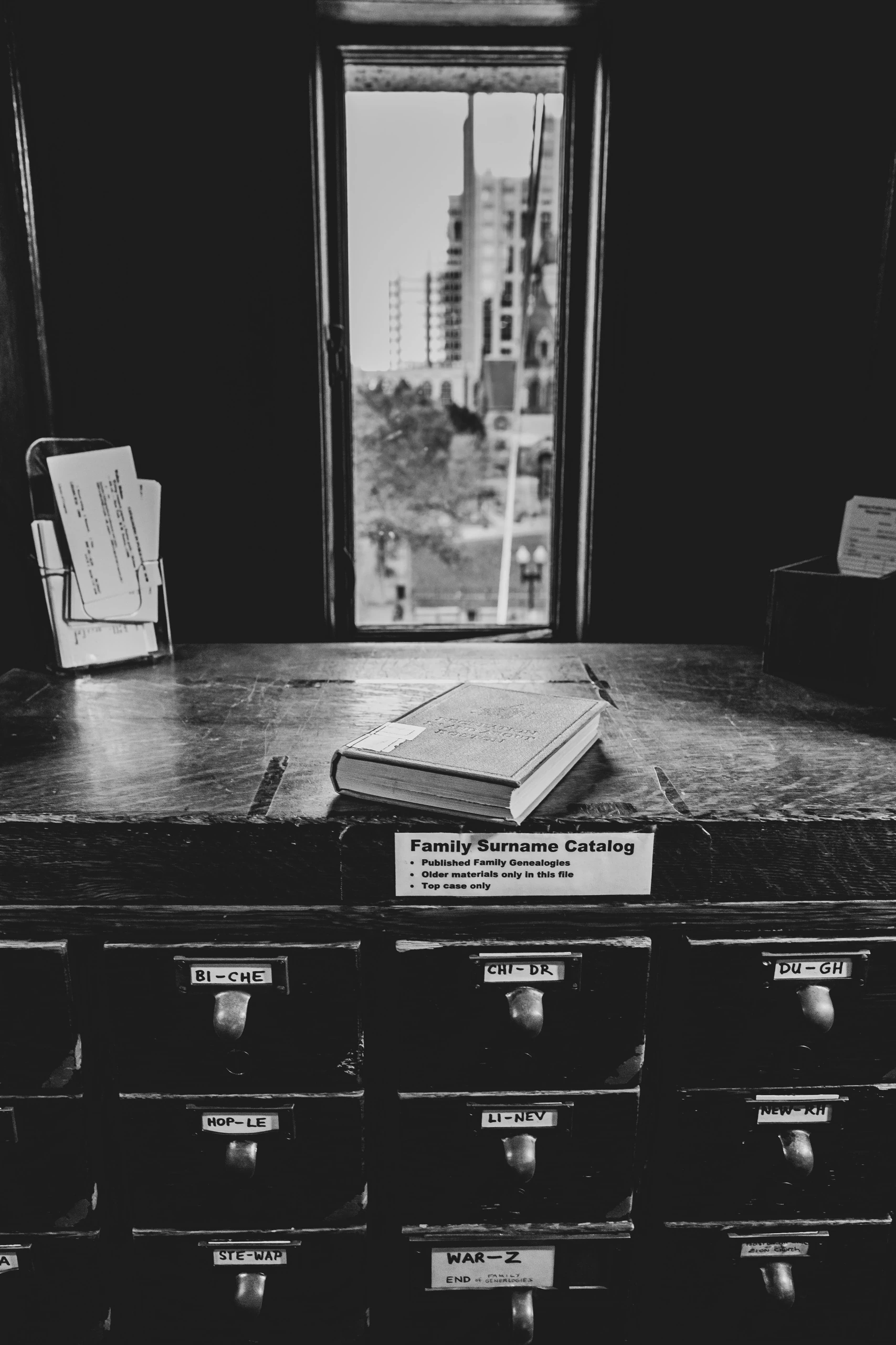 A wooden card catalogue with a notebook on top, in front of the window onto the city.