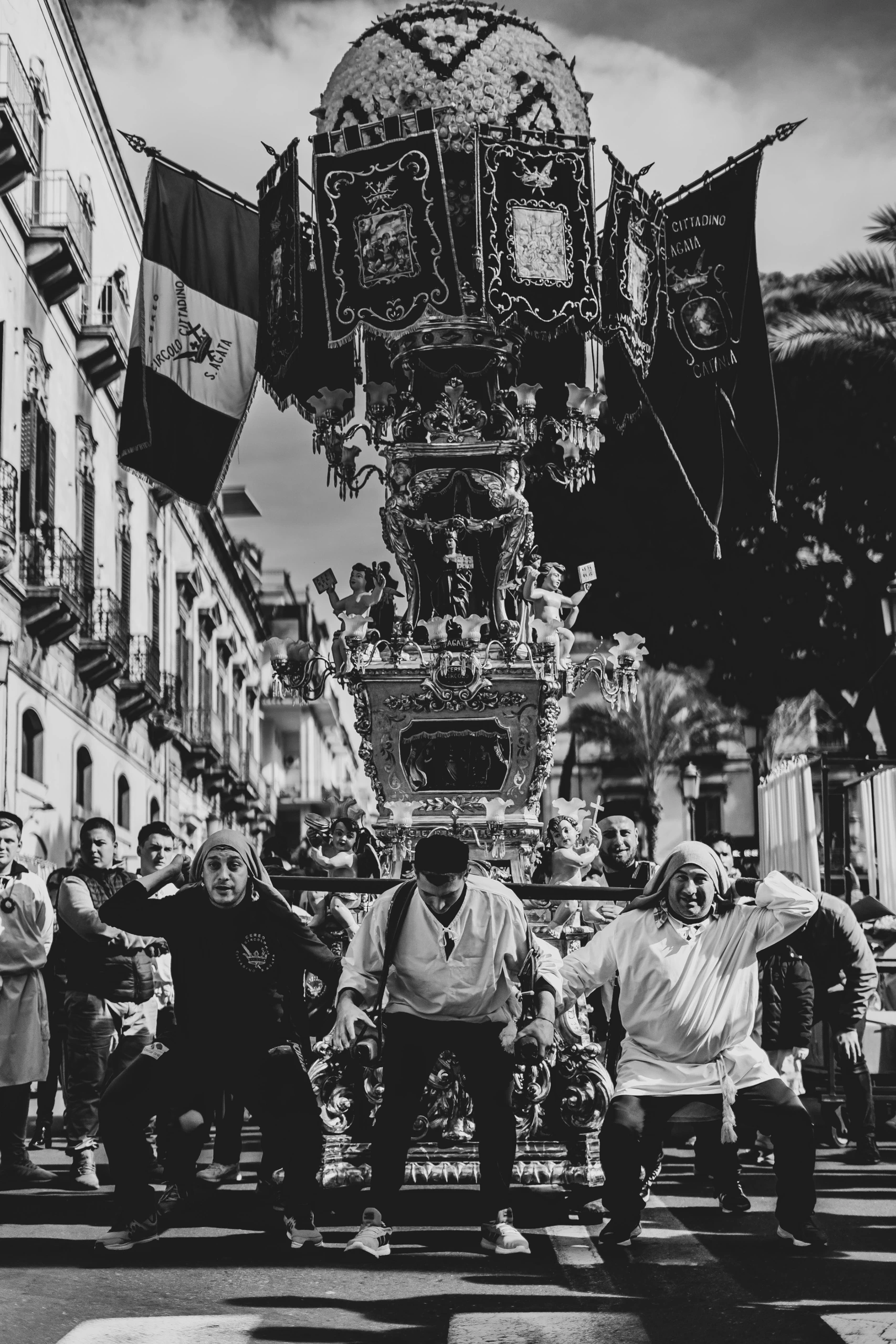 A candelora carried on shoulders along the street, among the flags.