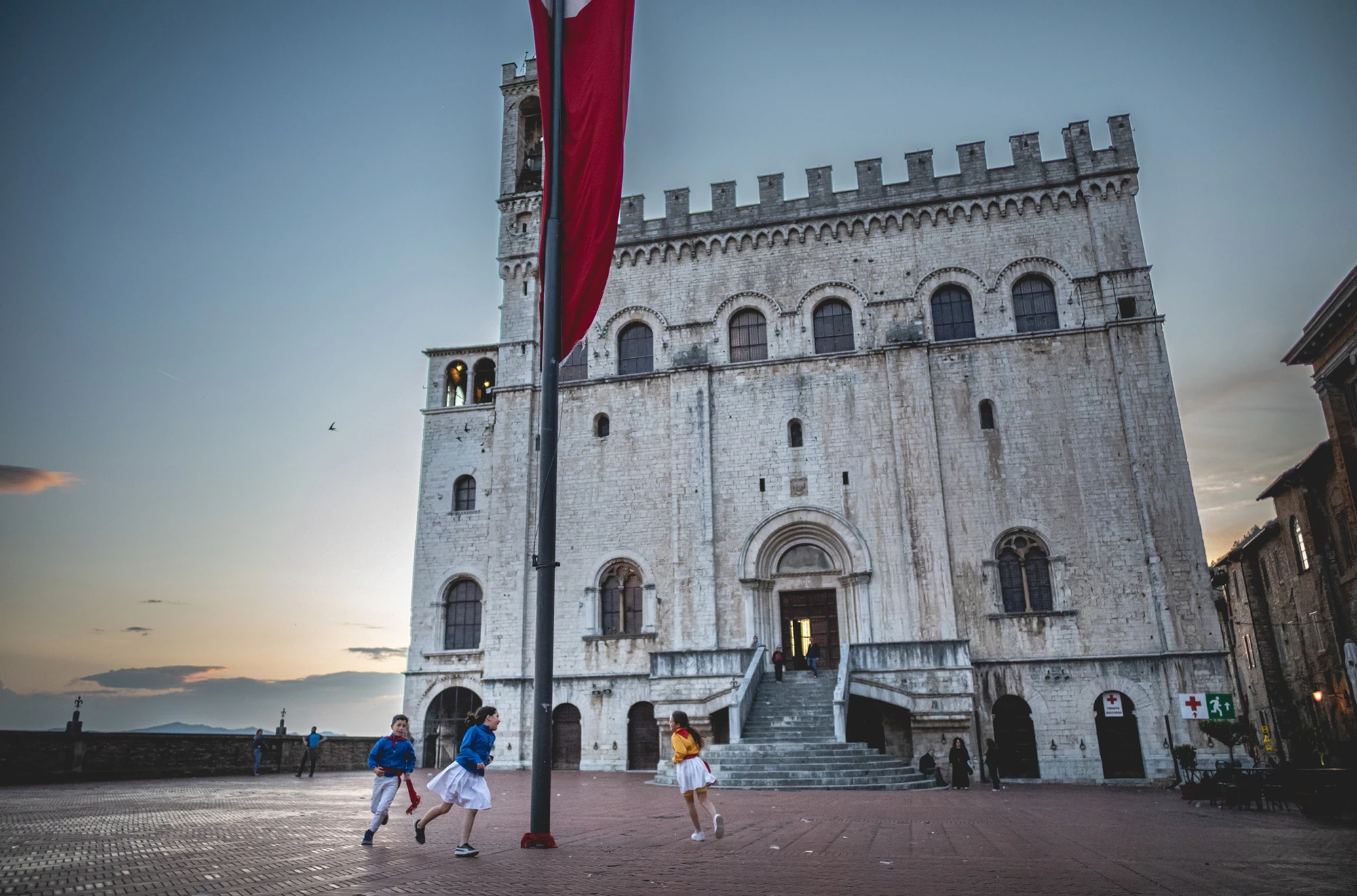 At dusk, the Palazzo dei Consoli with its red banner and children running in the square.
