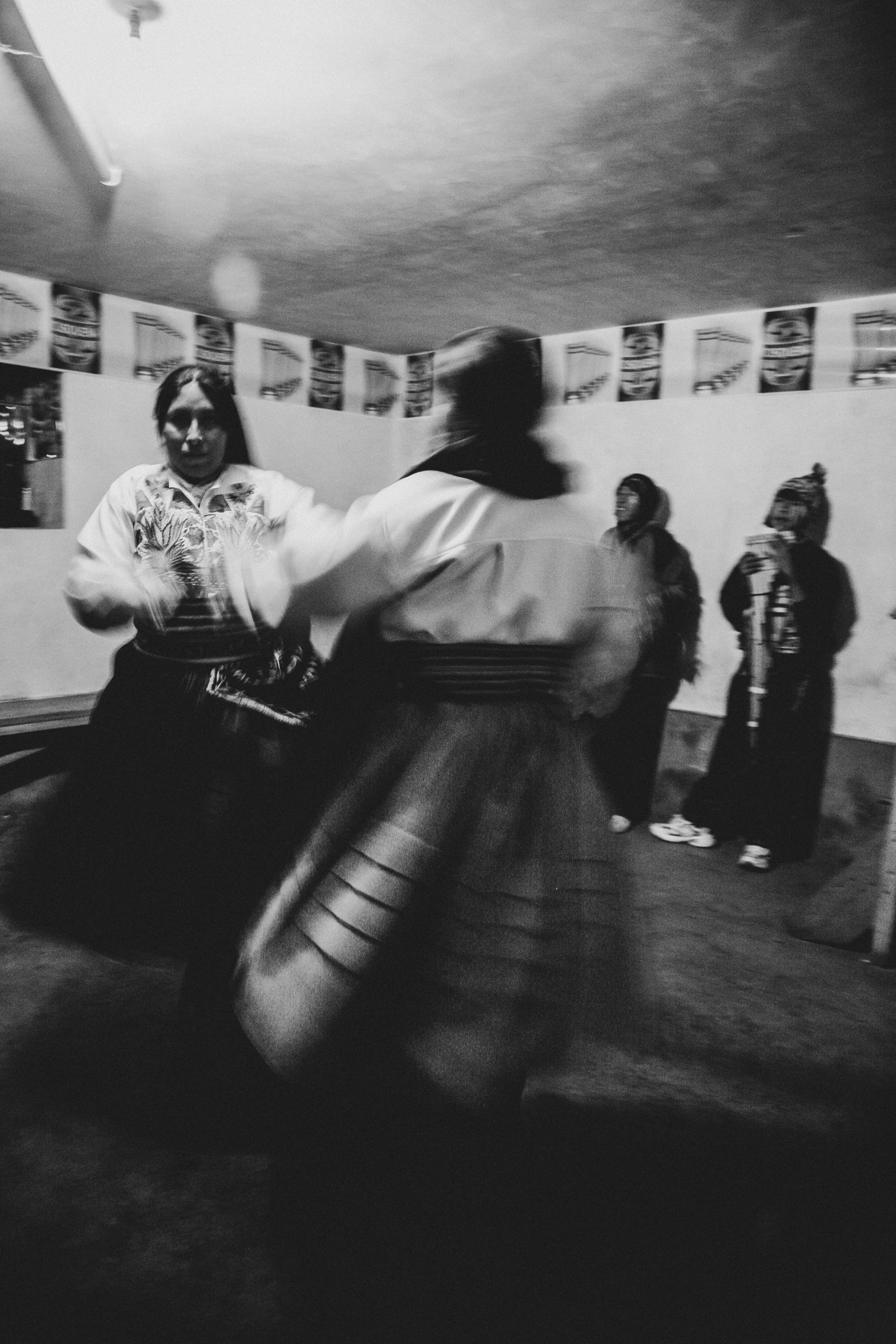 Indoors, two women in traditional skirts dance in a blur; others stand along the wall.