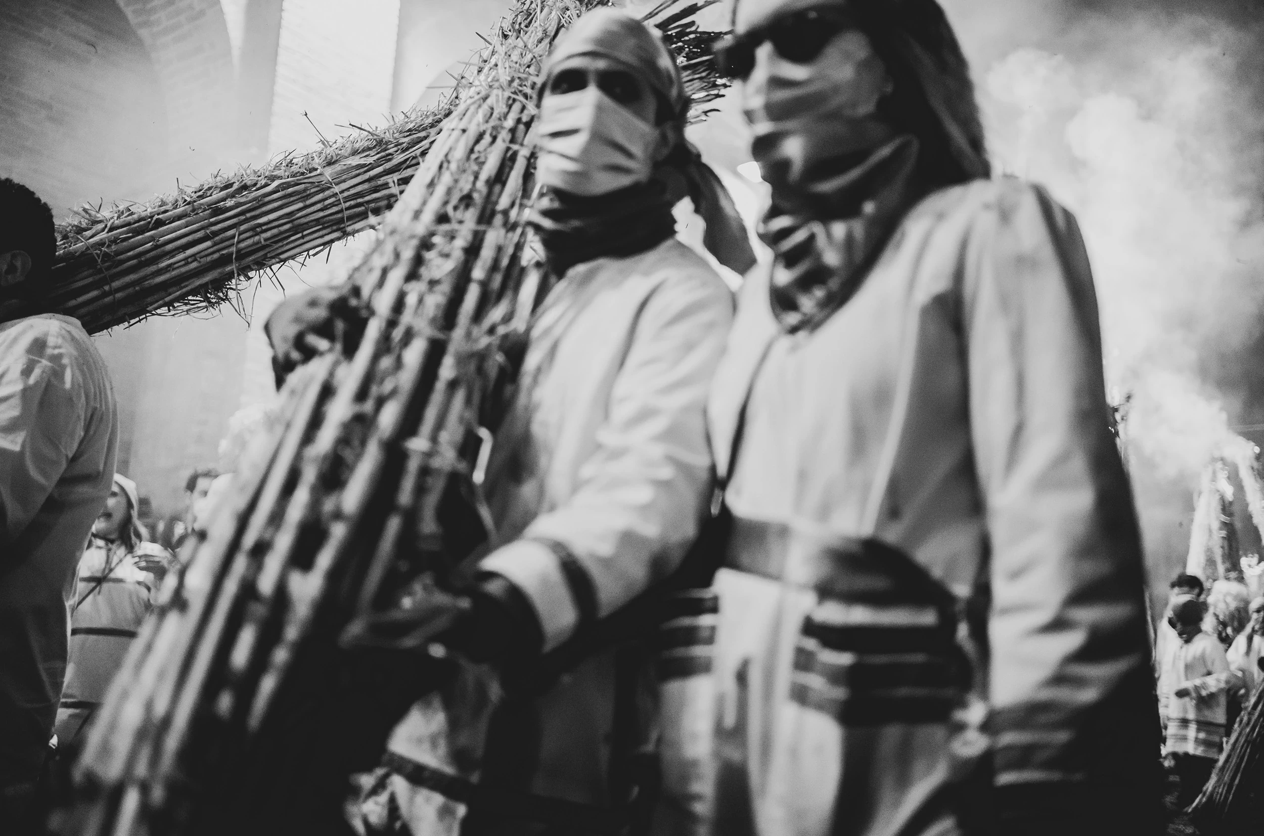 Two figures with bound faces carry bundles of cane on their shoulders, in the smoke.
