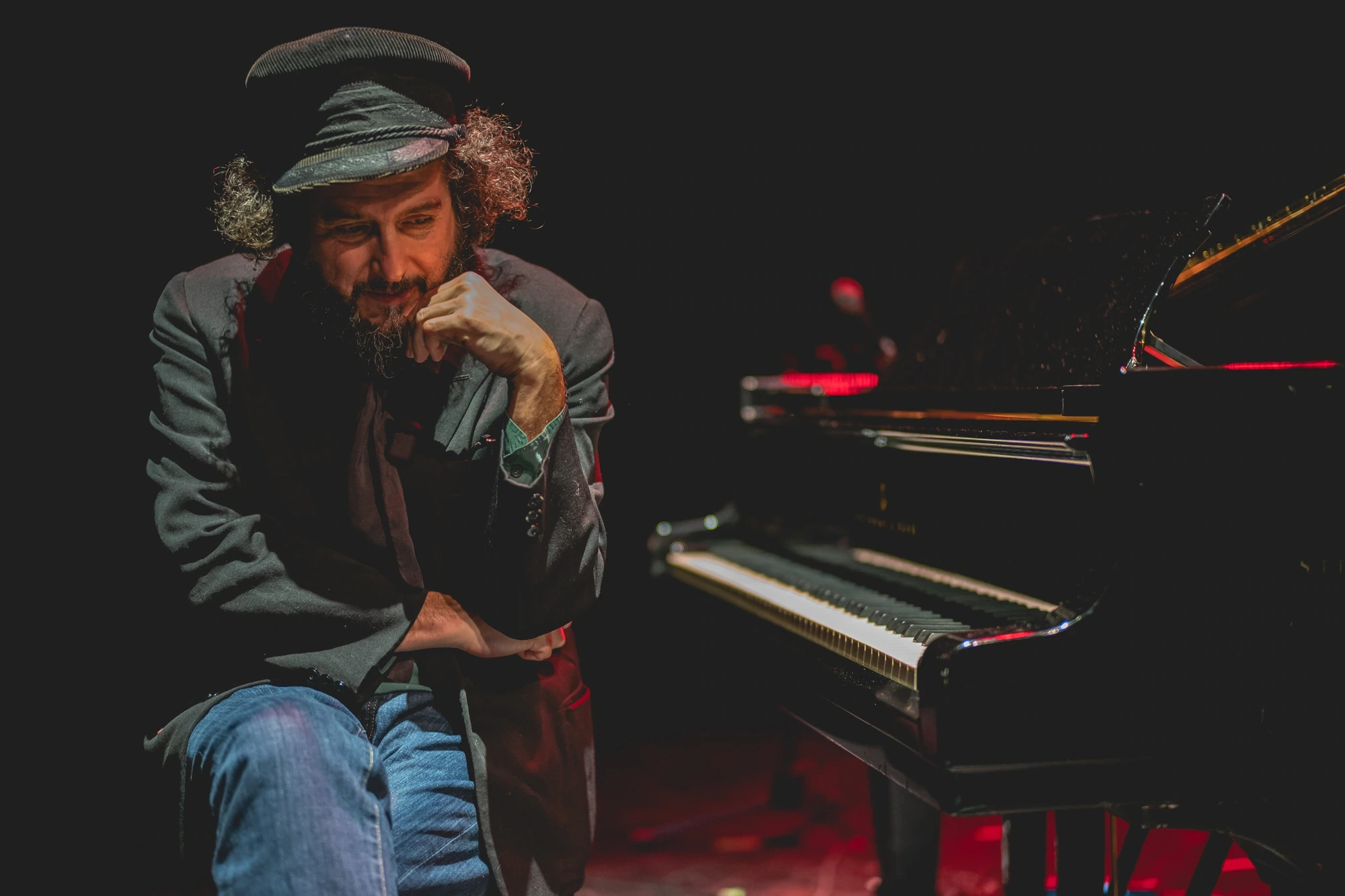 At the grand piano, hand on chin and cap on head, in a pause.