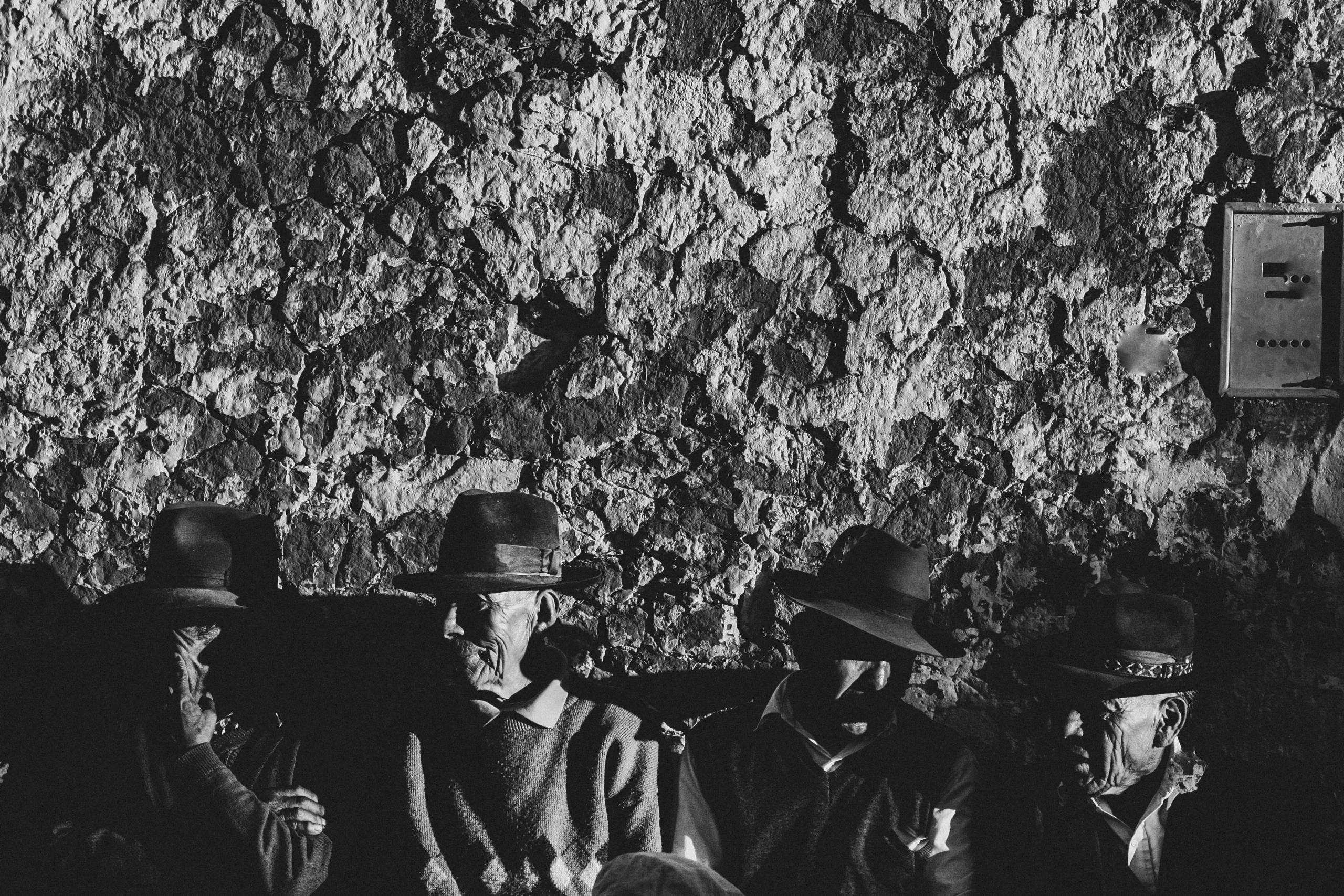 Men in hats sitting beneath a rough stone wall.