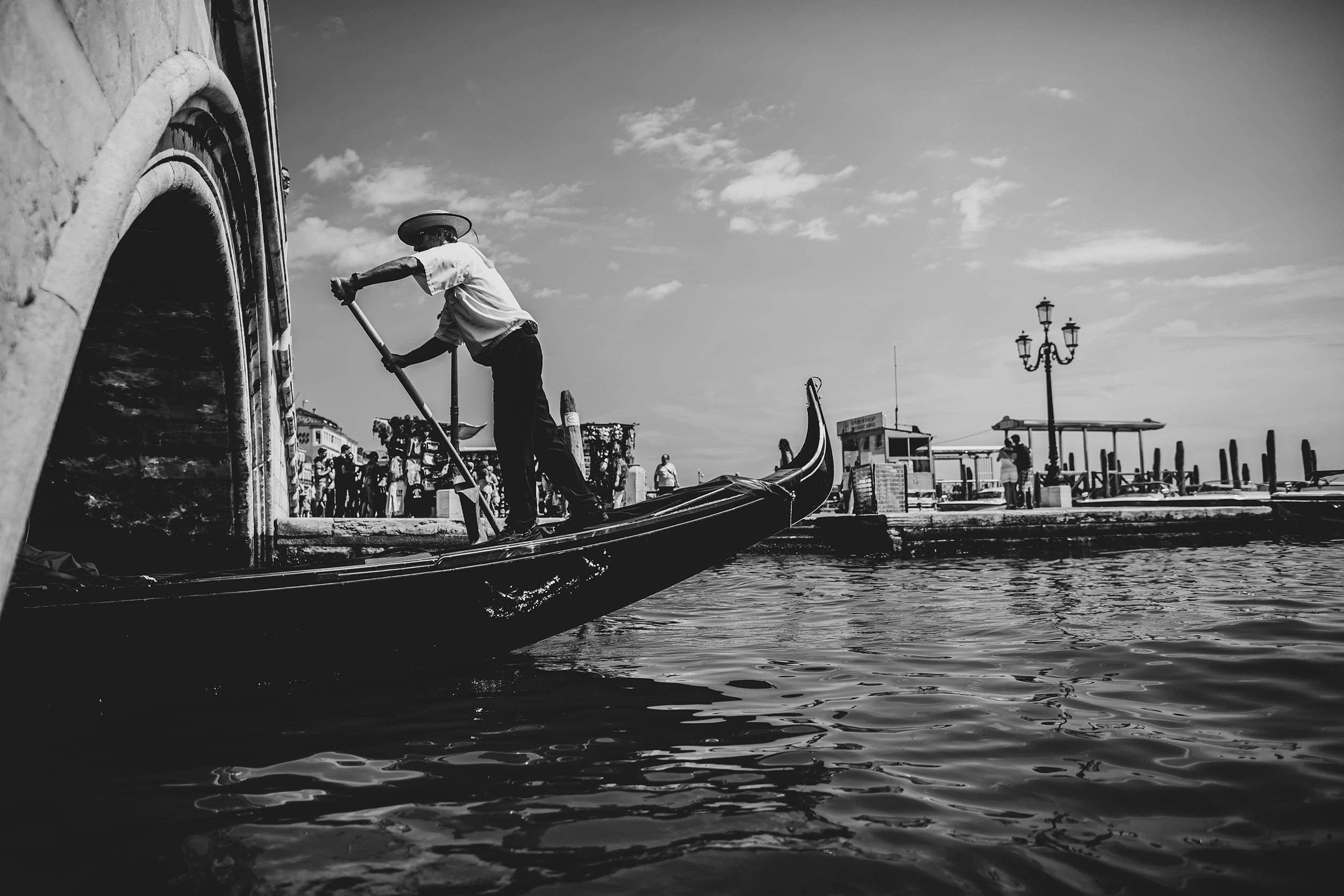 From under a bridge arch, a gondolier rows towards a crowded embankment.