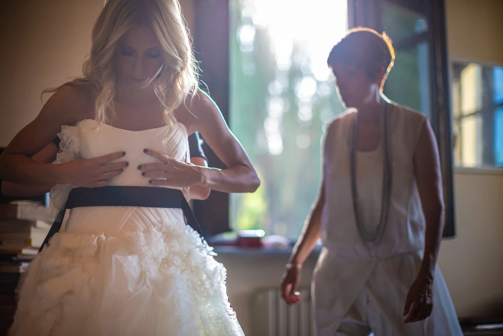 The bride adjusts her dress at the window, helped by a woman.