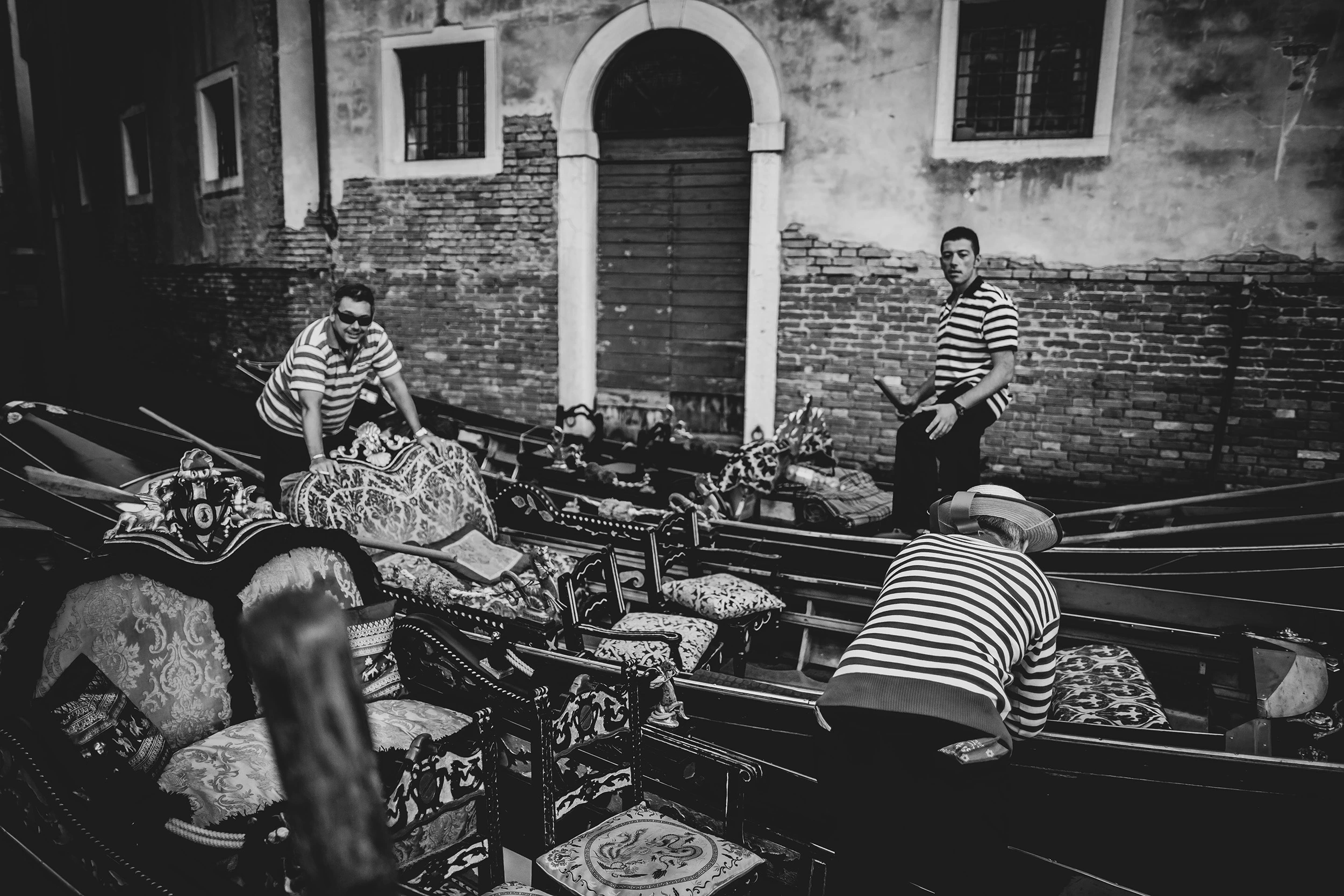 In a narrow canal between brick walls, three gondoliers around moored gondolas and embroidered cushions.