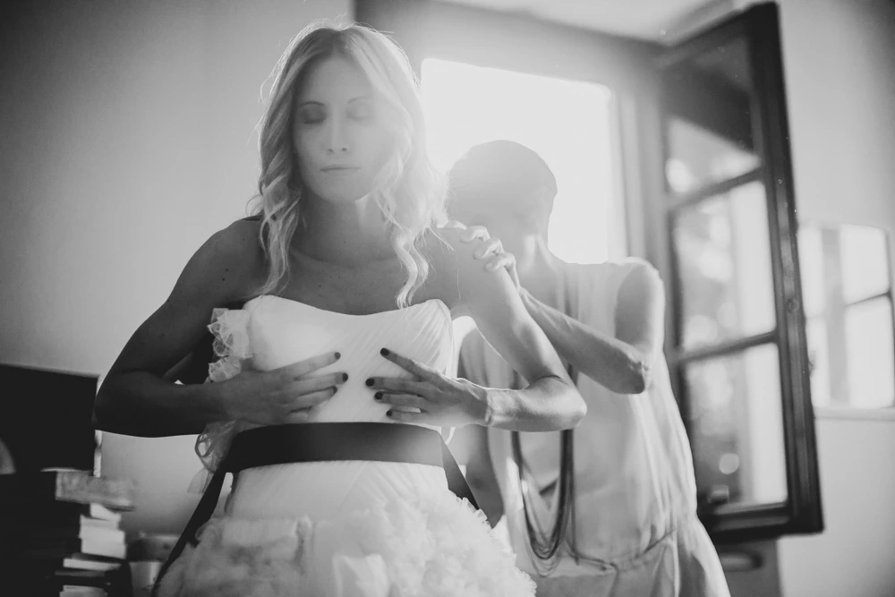 At the window, a woman fastens the dress at the bride's back.