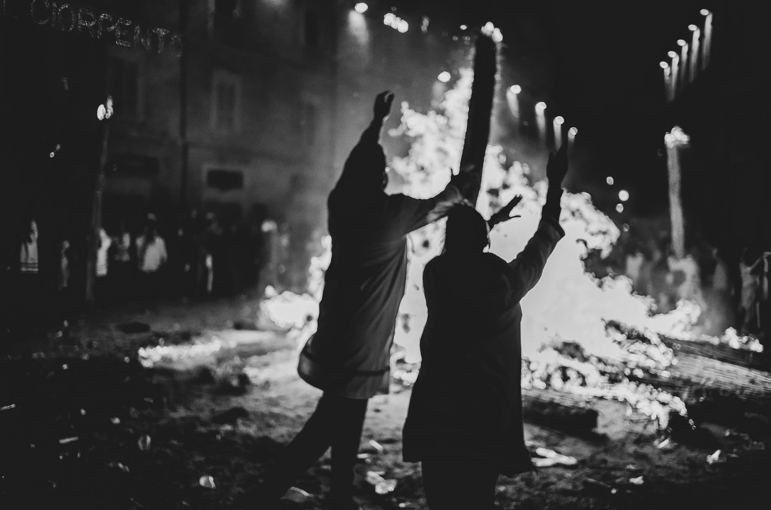 In front of the bonfire, two figures raise their arms.