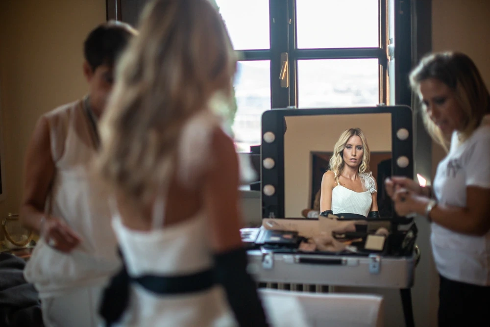 From behind at the lit mirror, while the make-up artist works.