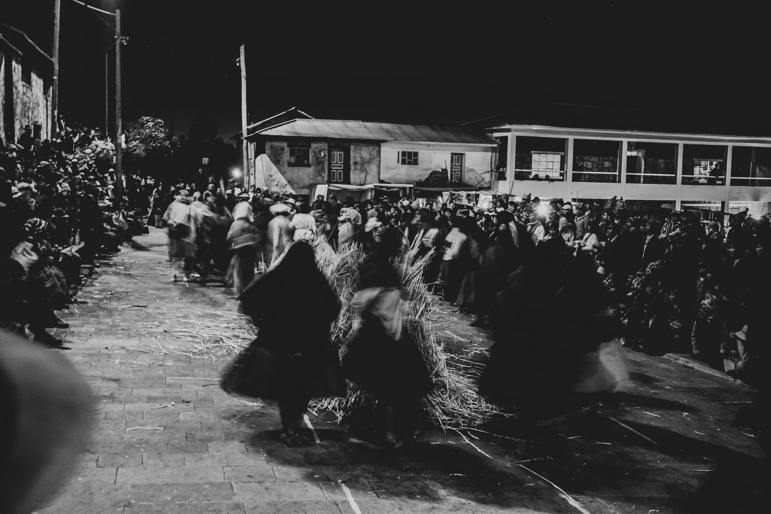 At night, blurred figures carry sheaves of grain along the street, in front of the crowd.