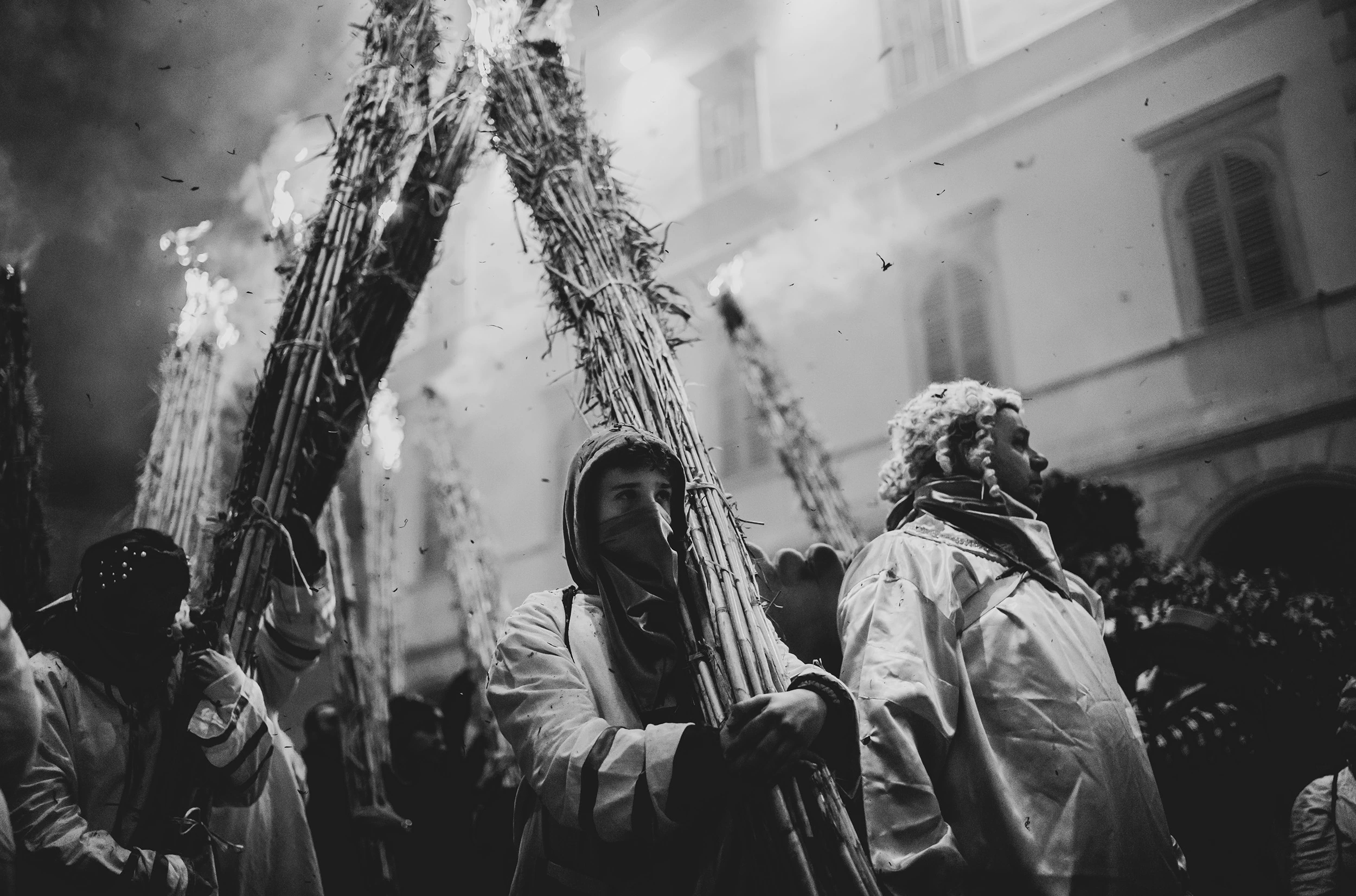 The bearers in white hold the lit bundles upright along the street.