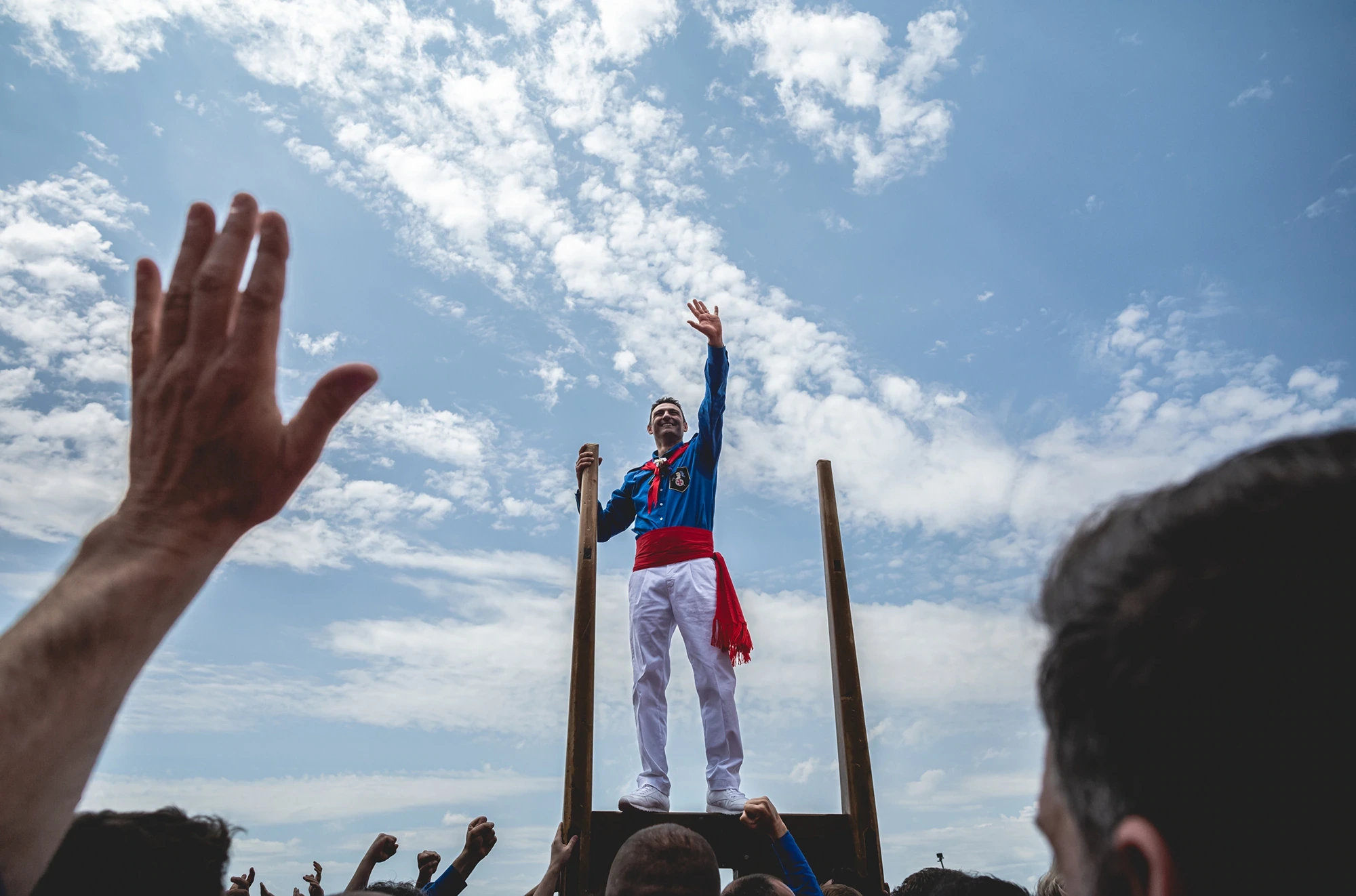 Atop the cero's shafts a man stands against the sky; in the foreground, the hands of the crowd.
