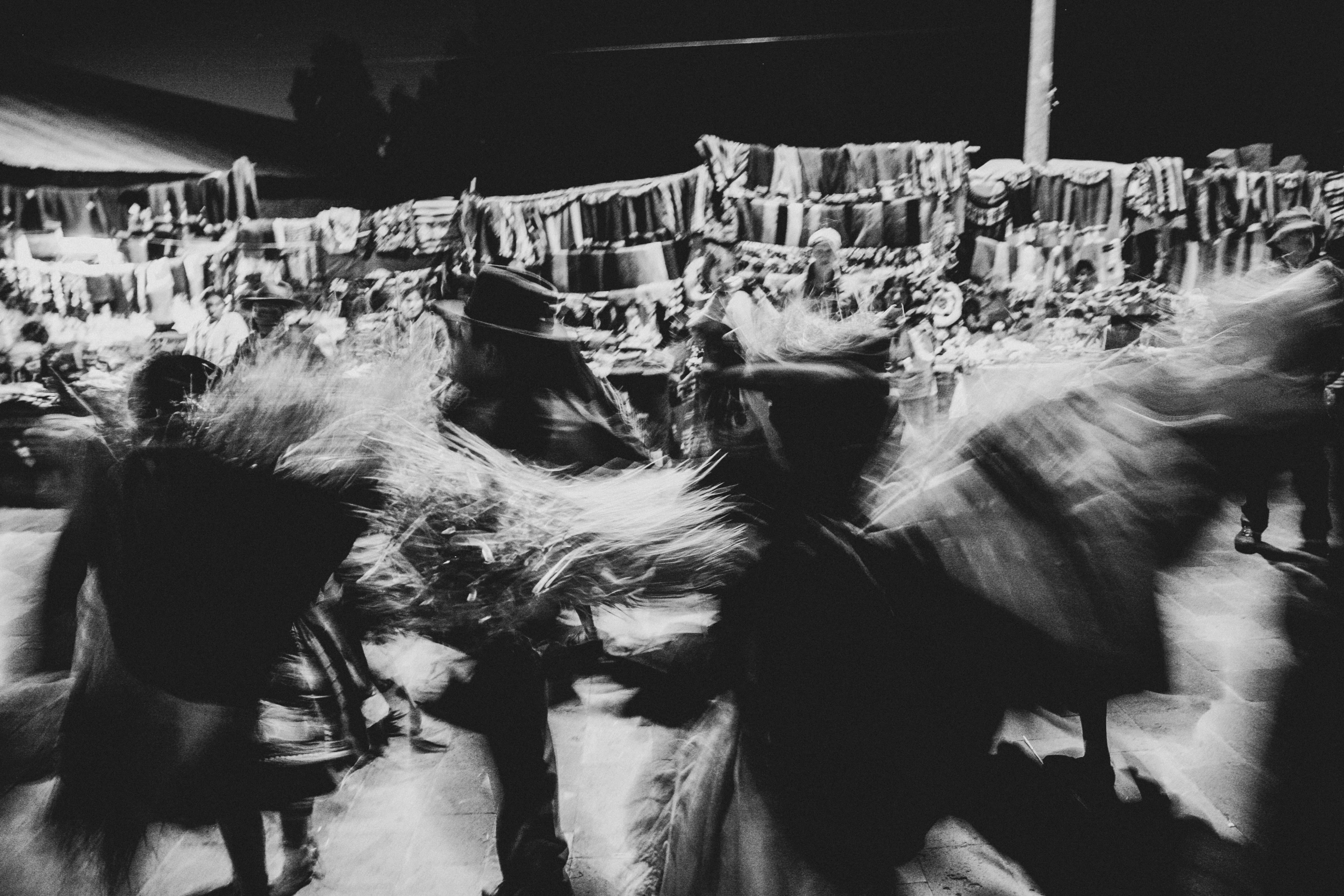 Dancers in fringed costume, blurred by movement, in front of the textile stalls.