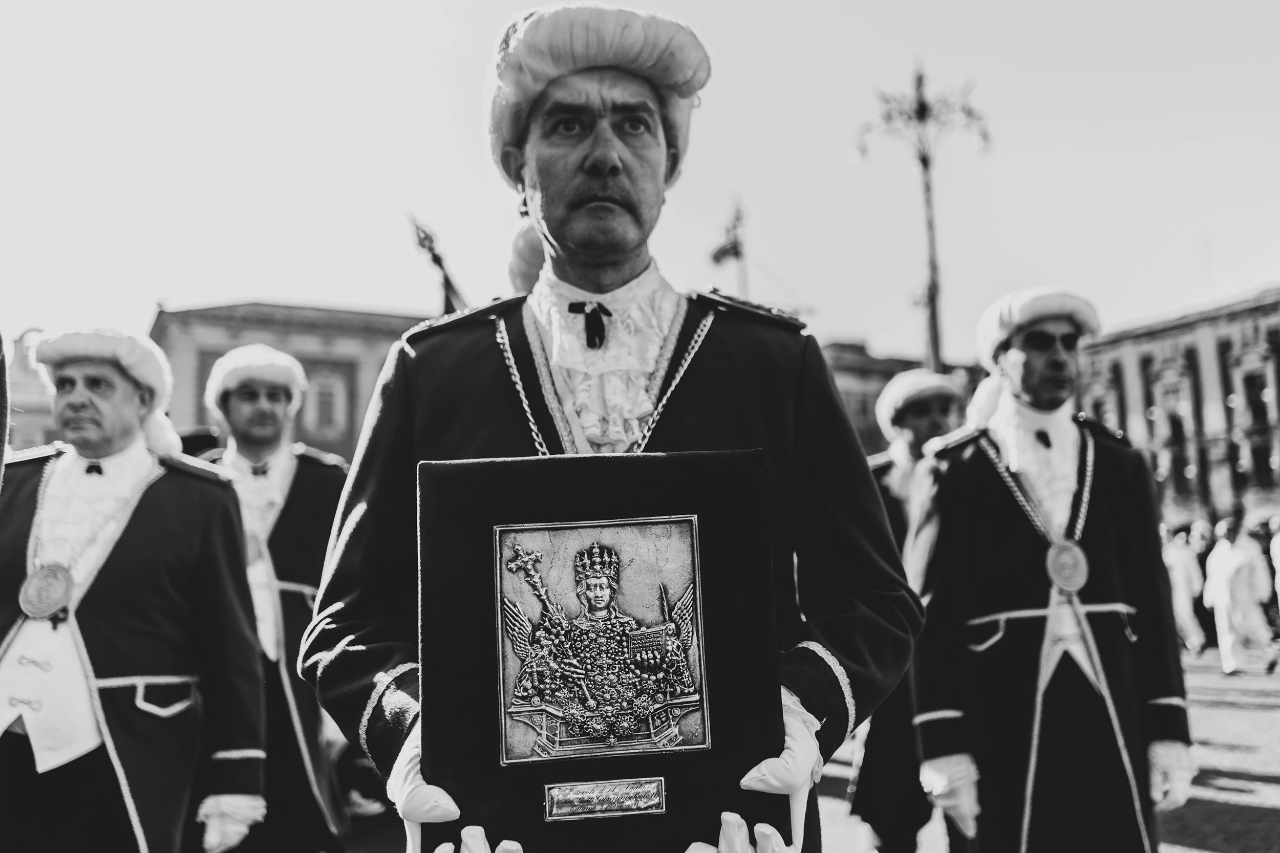 Brethren in ceremonial dress and white wigs; one holds the framed effigy of the saint.