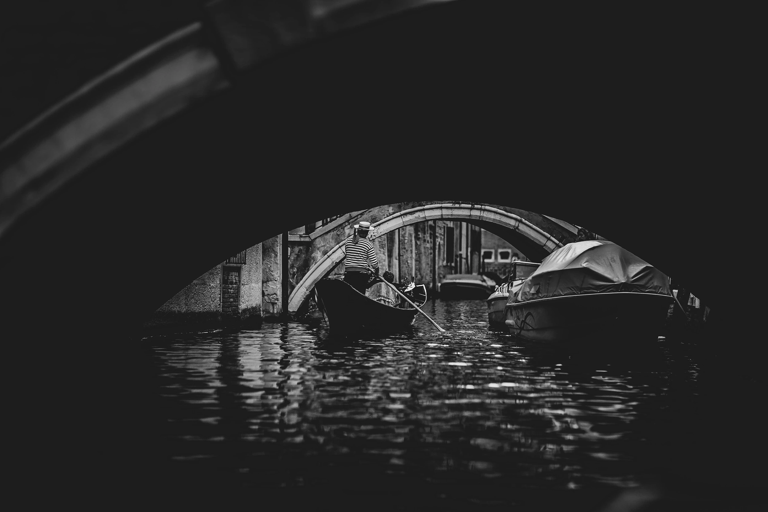 Under a dim bridge, moored boats and reflections on the water.