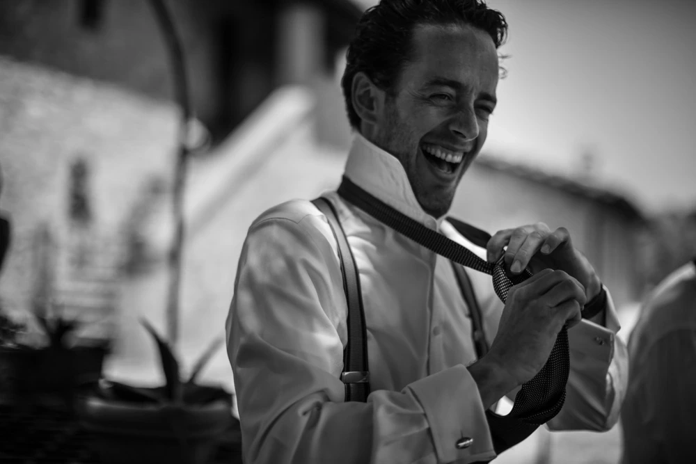 The groom laughs as he knots his tie, in braces.