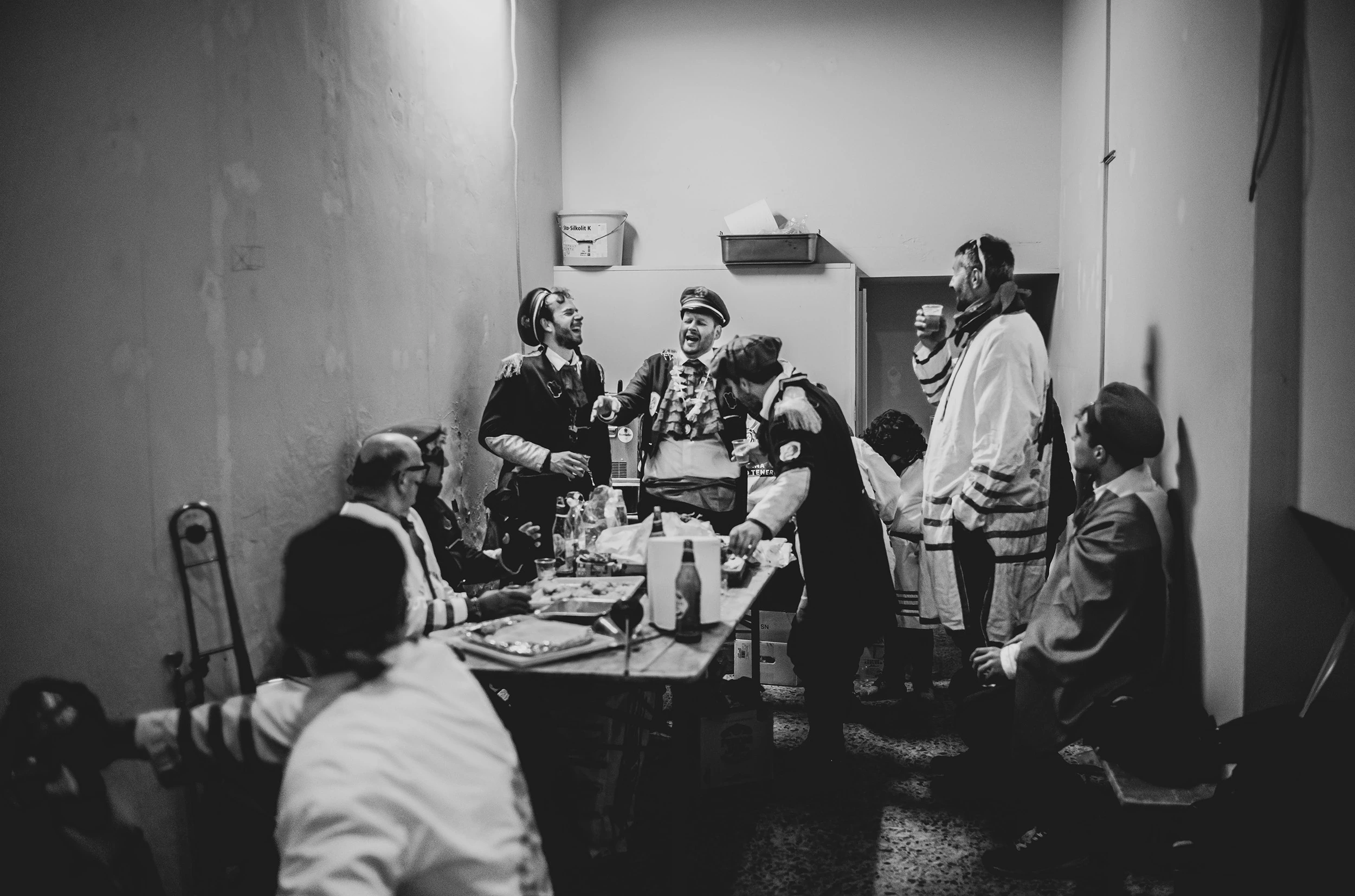 Indoors, the costumed participants at table among bottles and plates.