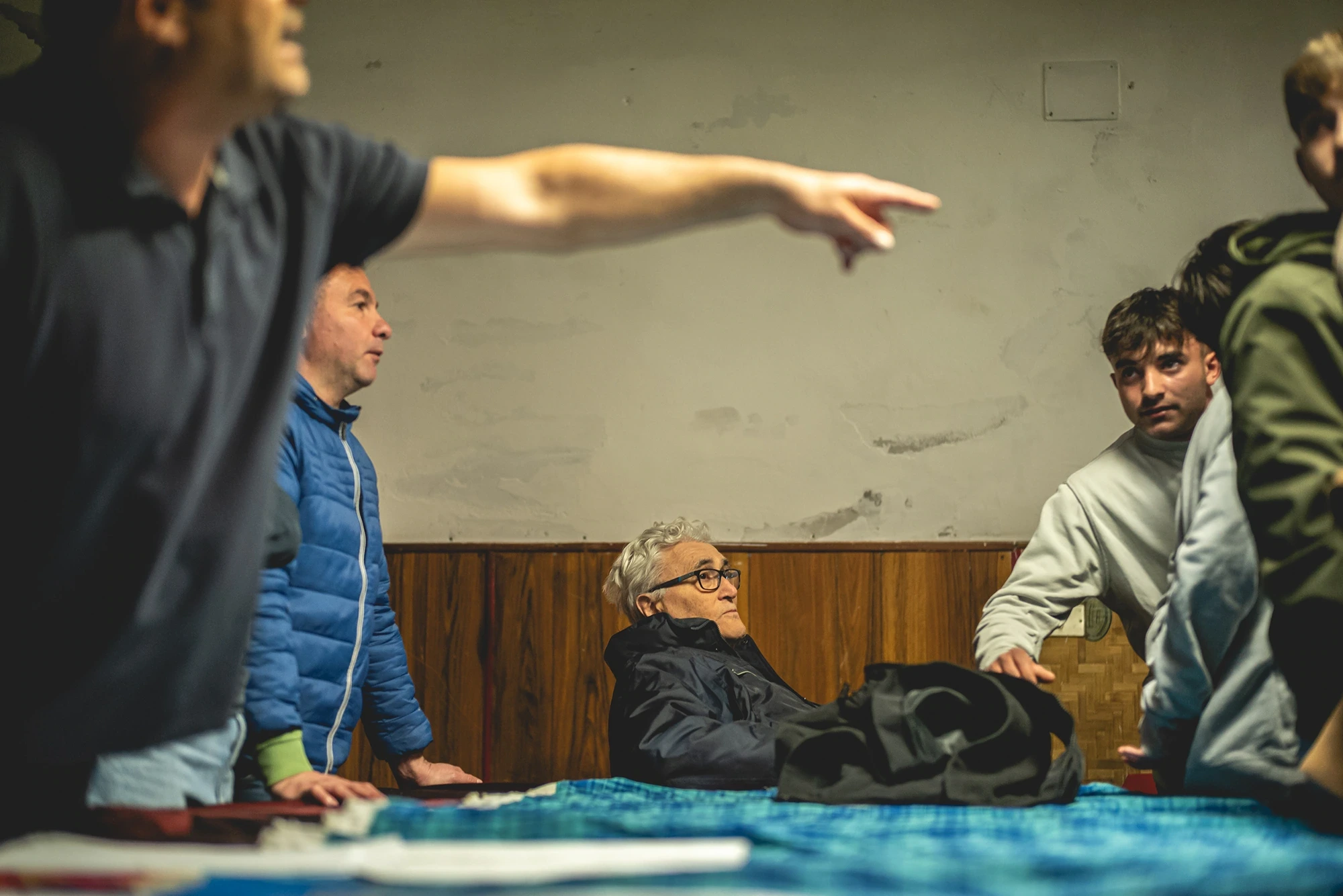 Indoors, men around a table; one points at something out of frame.