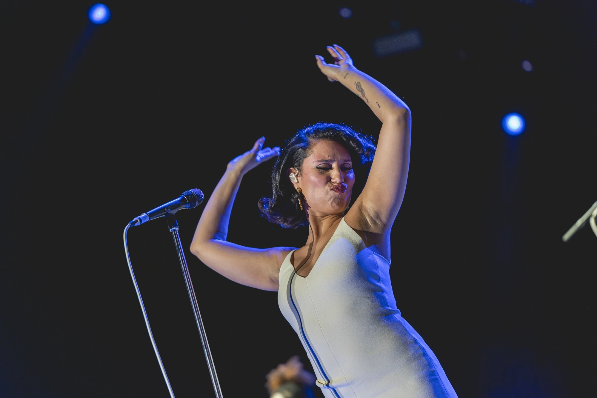 In a white dress and blue light, arms raised above the microphone.