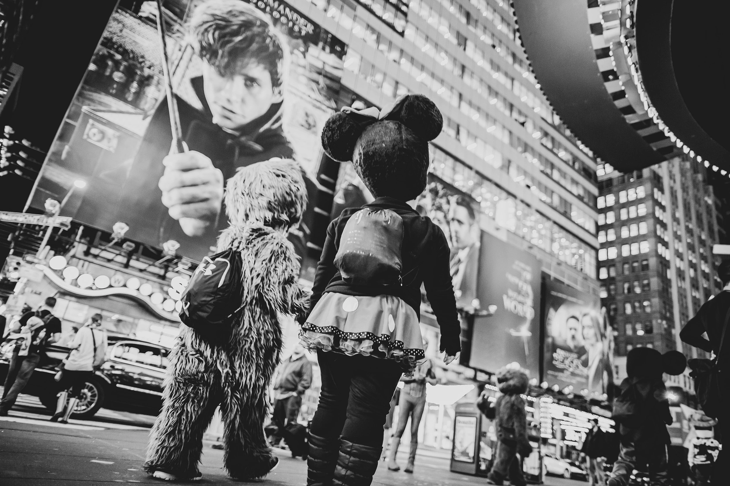 In Times Square, two costumed performers seen from behind beneath the billboards.