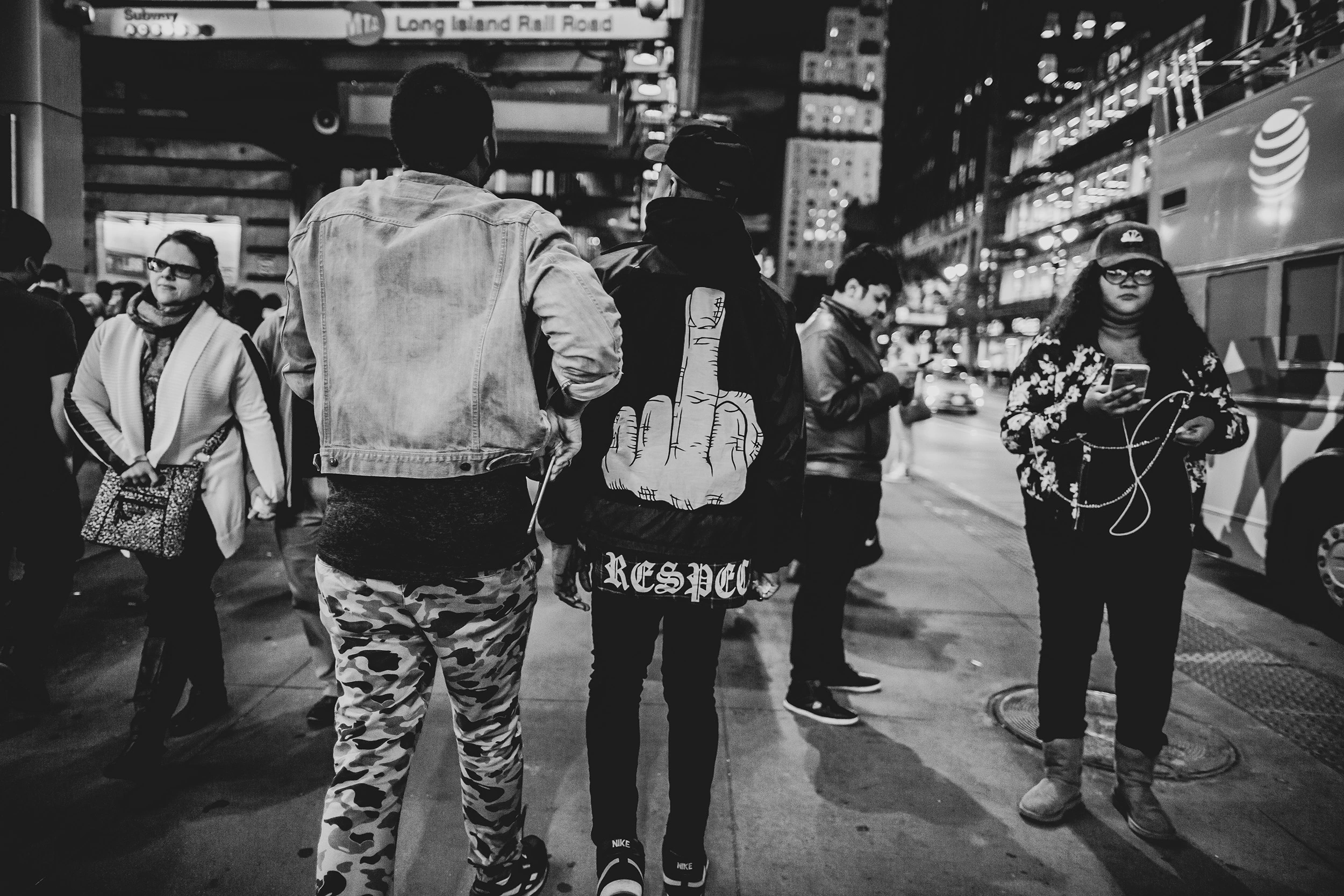 Passers-by from behind on a midtown street, beneath the illuminated signs.