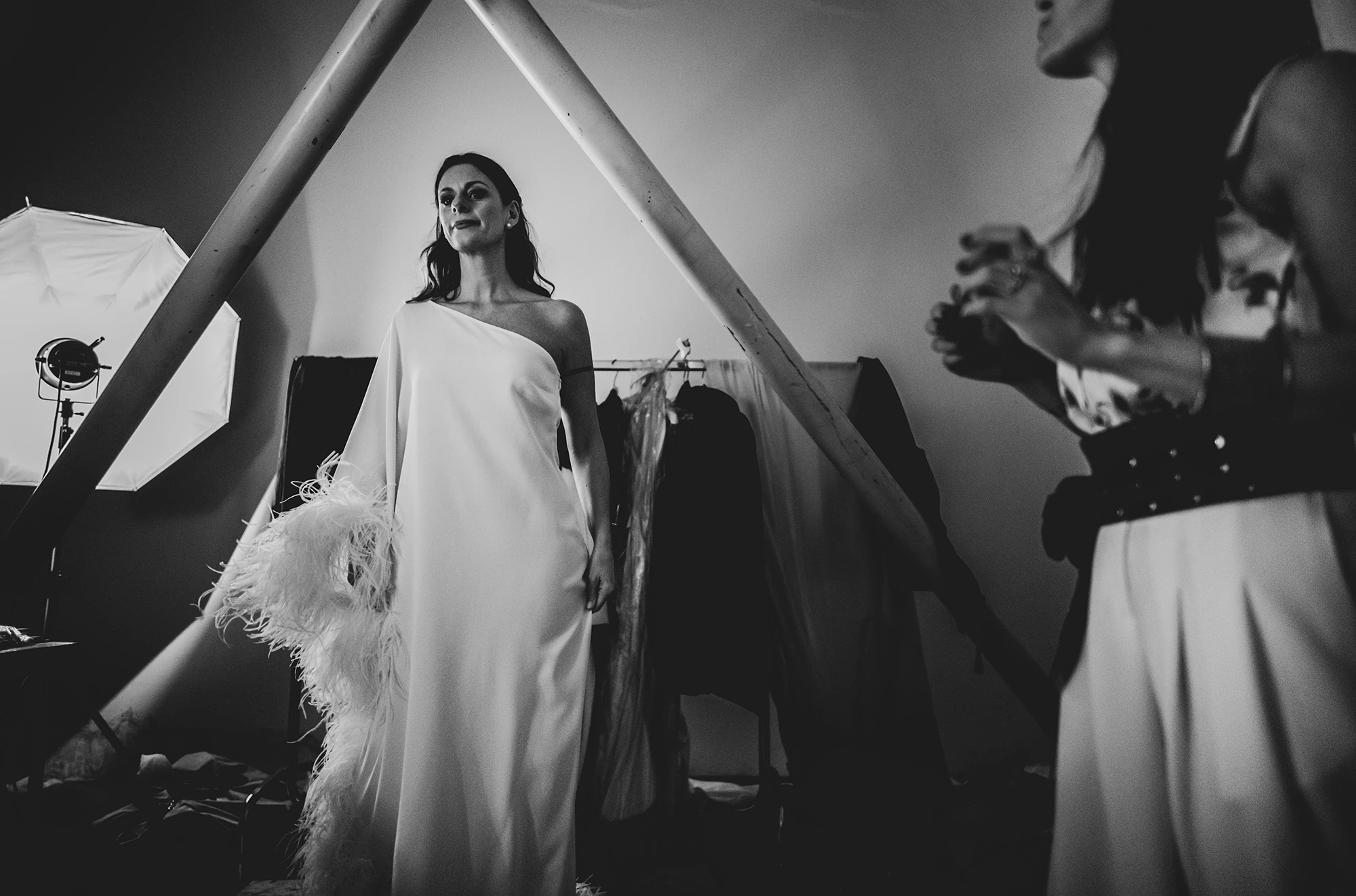 In a one-shoulder dress, standing in the lamp-lit room.