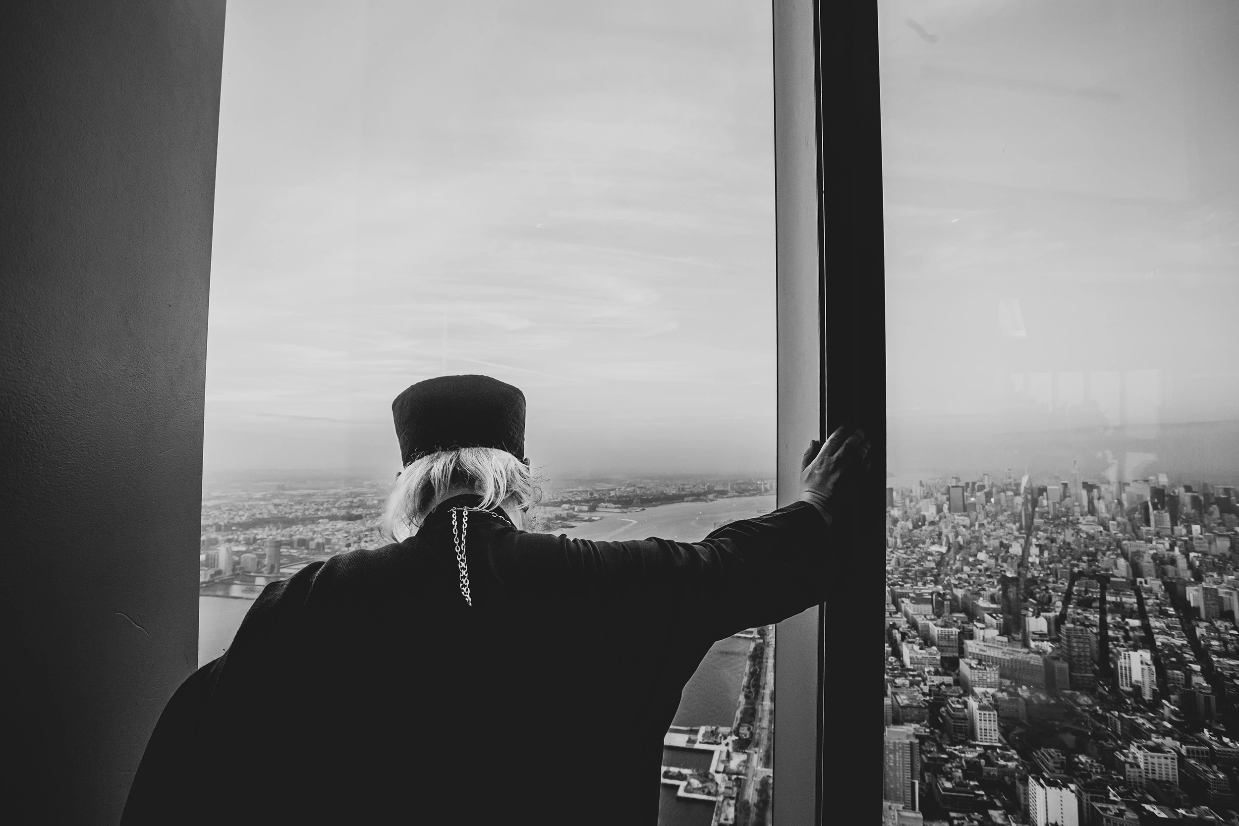 Back turned at a high window, a person looks down over the city.