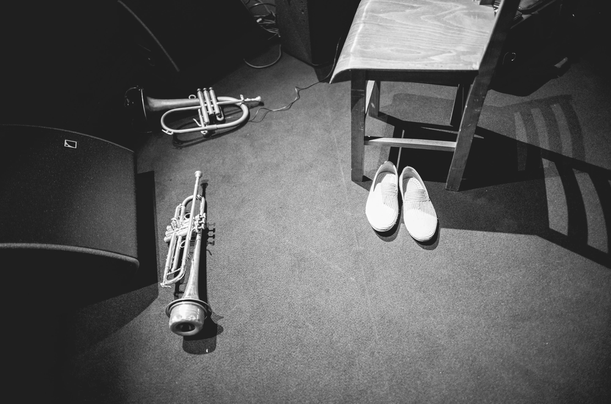 A trumpet set down on the floor beside a chair and a pair of white shoes.