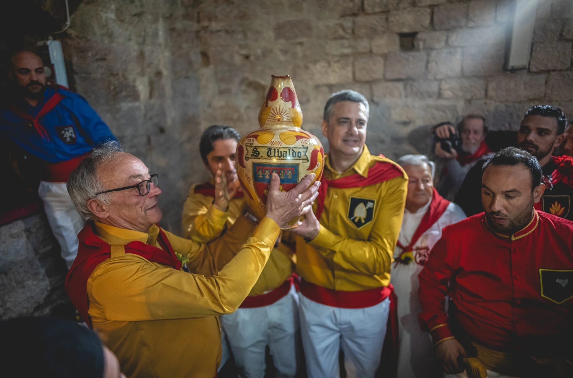 Men in yellow and red shirts raise a small ceramic amphora.