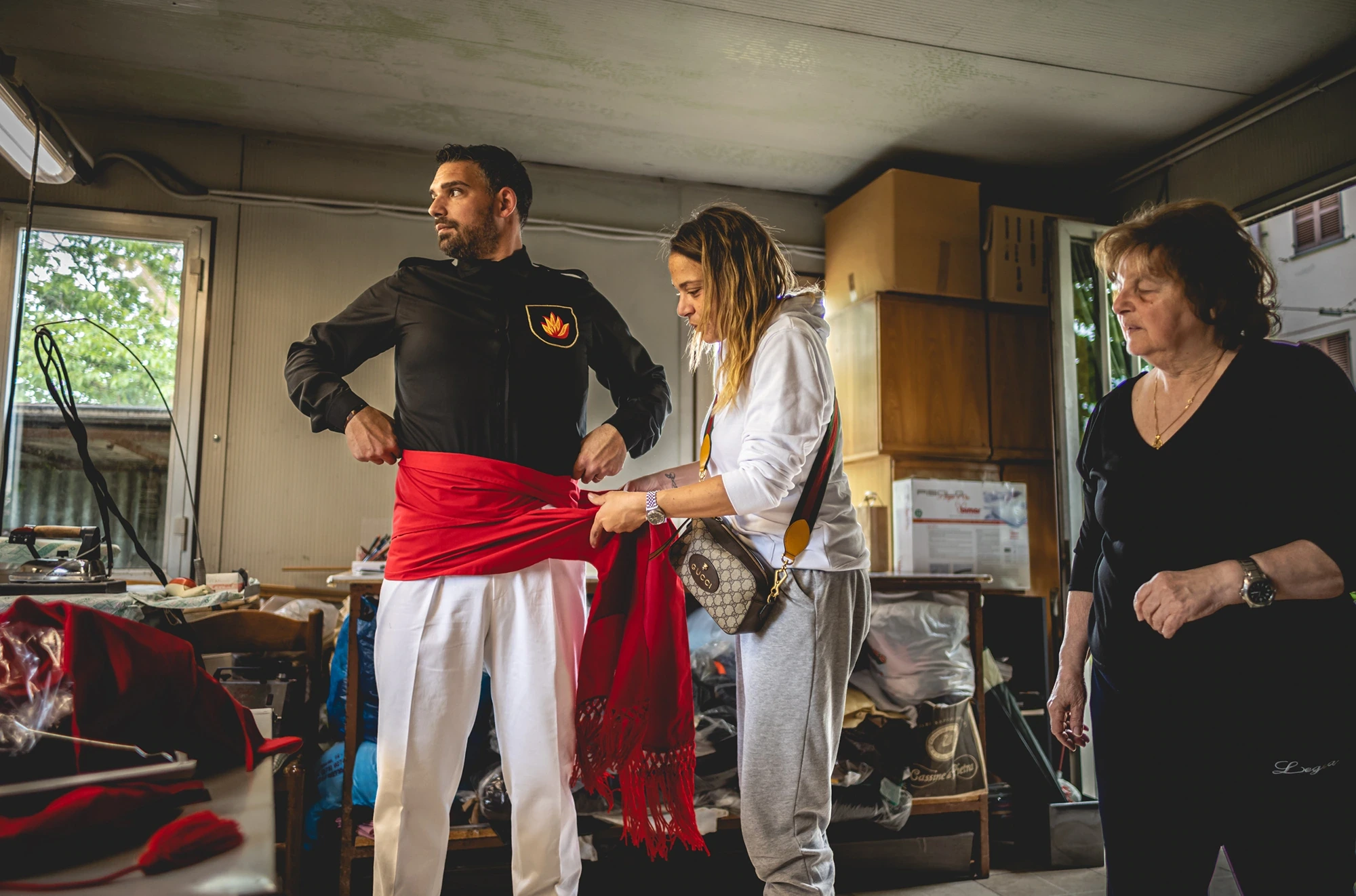 At home, a woman knots the red sash at the waist of a ceraiolo in a black shirt.