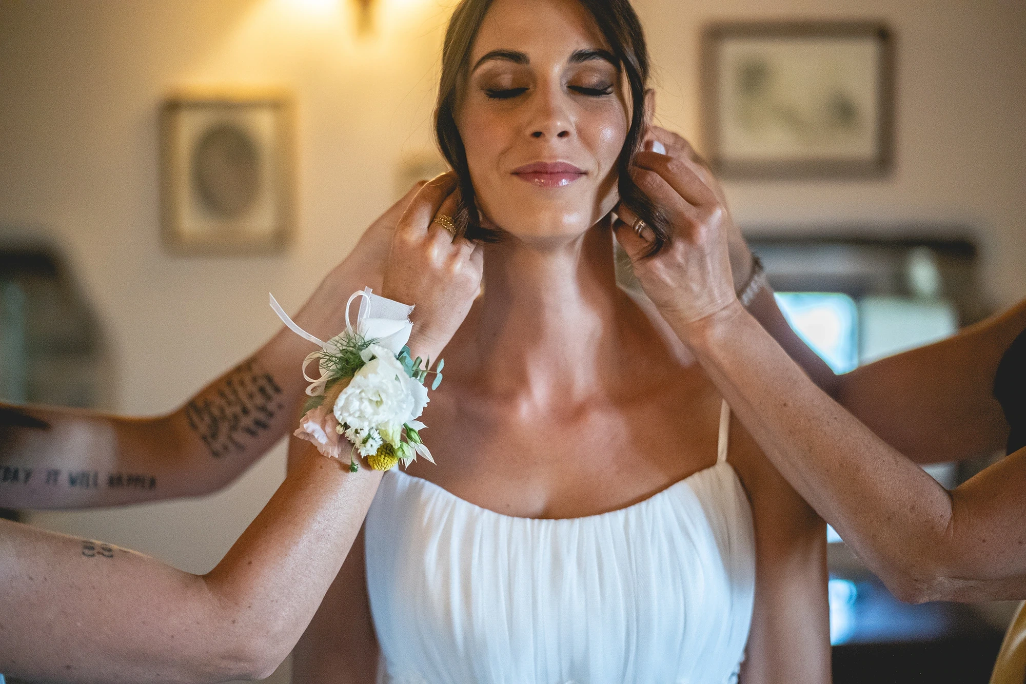 Hands fasten the necklace; on her wrist, the small corsage.