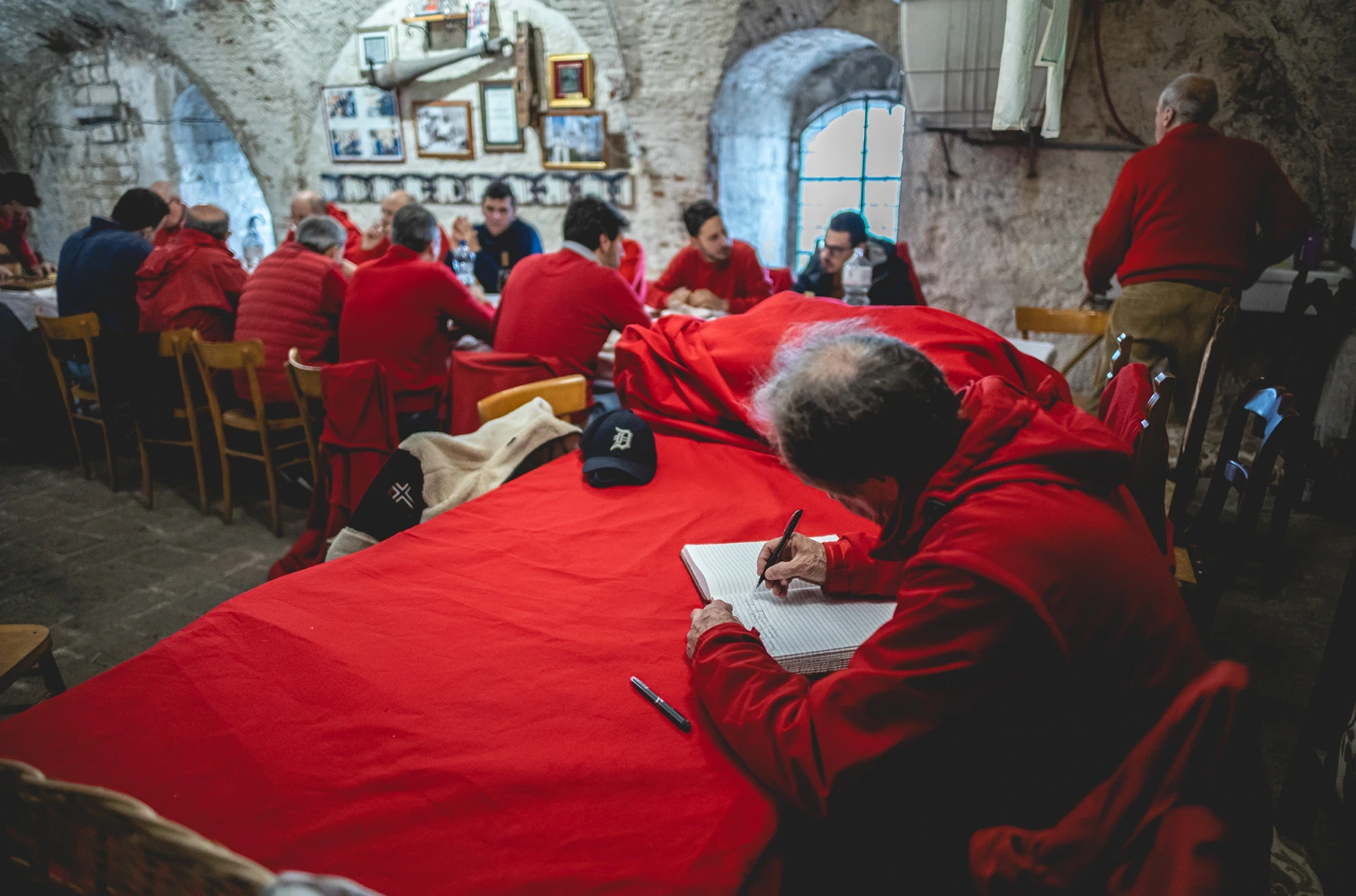 In a vaulted room, men in red jackets at cloth-covered tables; one is writing.