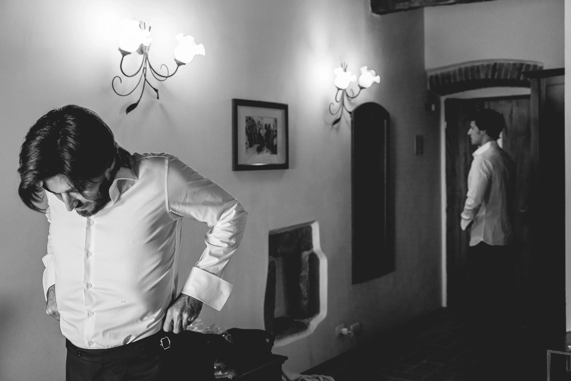 The groom tucks in his shirt; beyond the door, another checks the mirror.