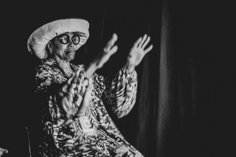 In a white beret and patterned coat, hands open in song.