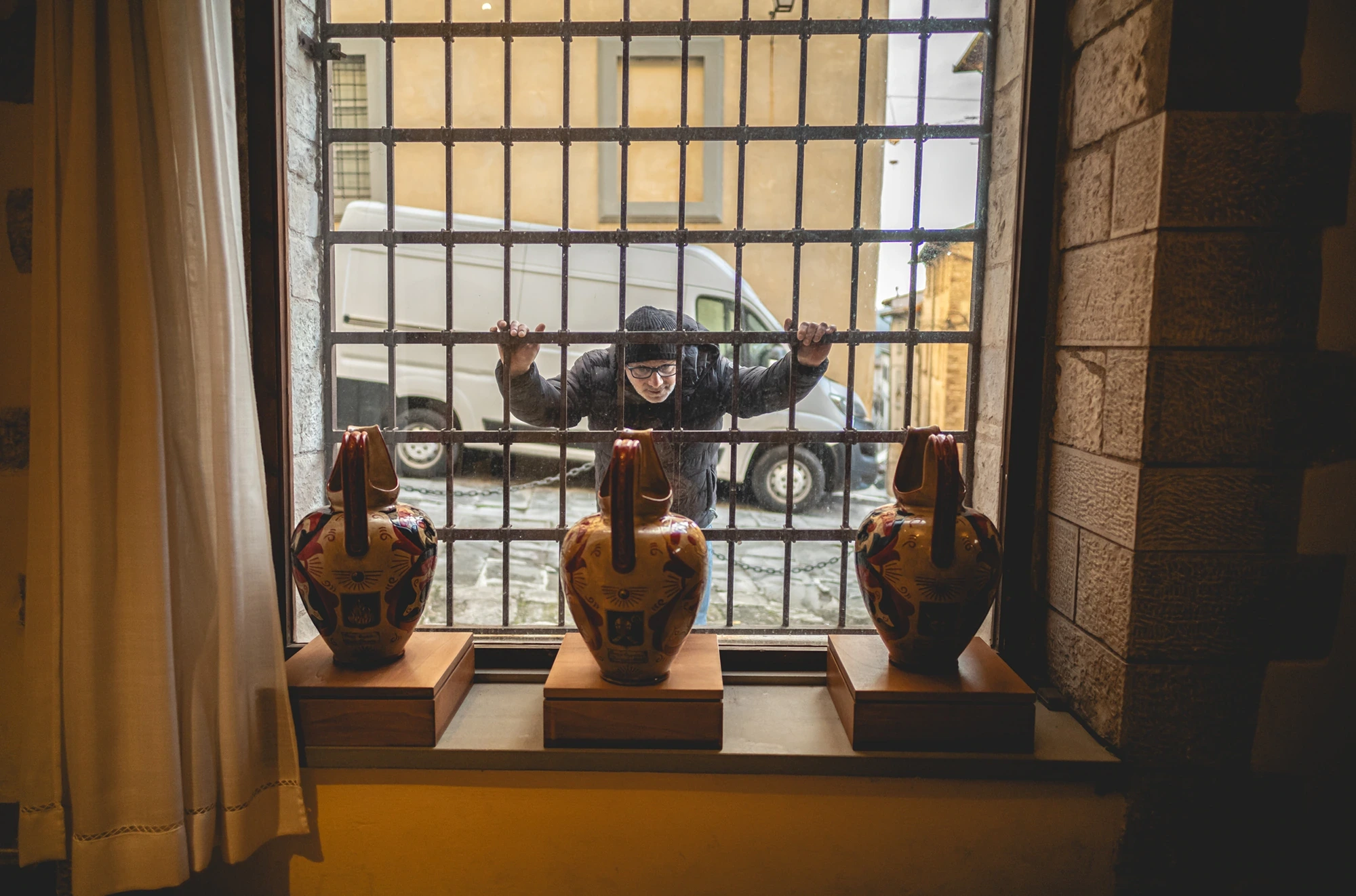 Three ceramic amphorae on the sill behind the grille; outside, a man gestures.