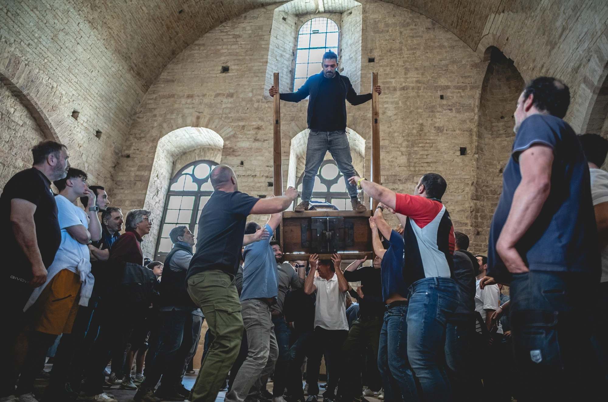 Inside a vaulted hall, a man stands on the wooden frame with open arms, held up by the group.
