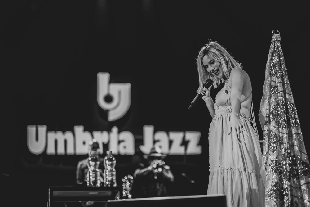 At the microphone in a pale dress, beside the festival sign.