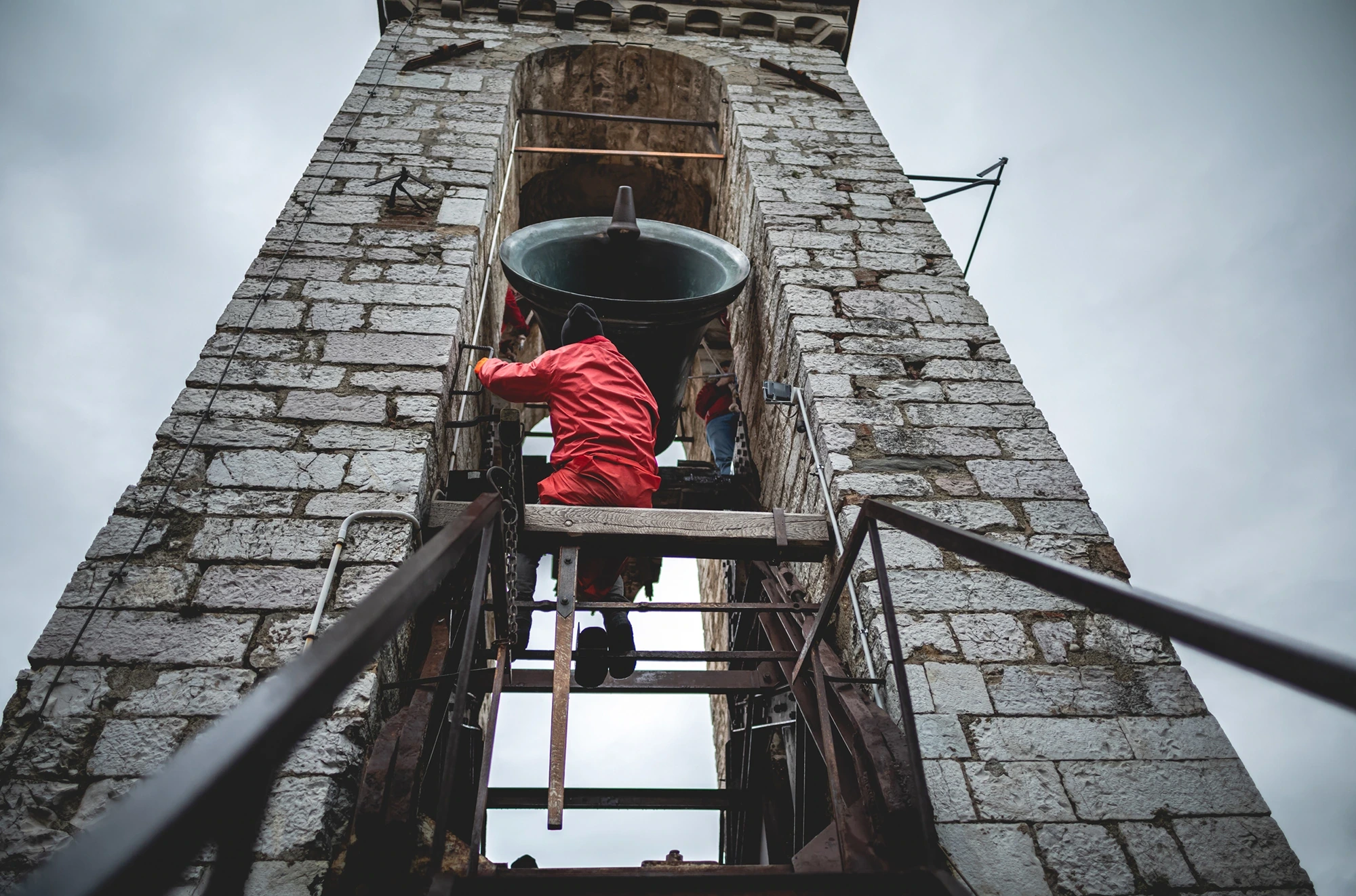 Inside the bell tower, a man in a red jacket climbs the ladder towards the bell.