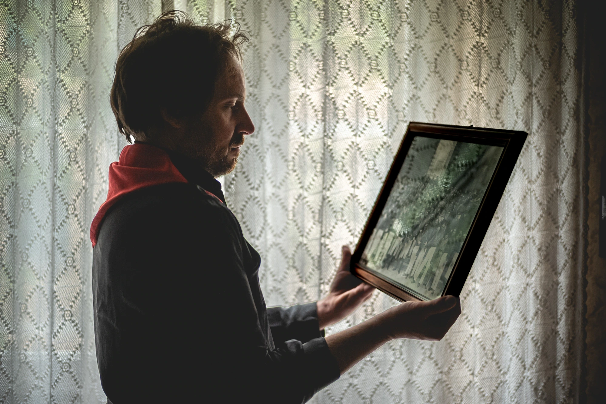 A man with a red sash holds a framed photograph, beside the curtain.