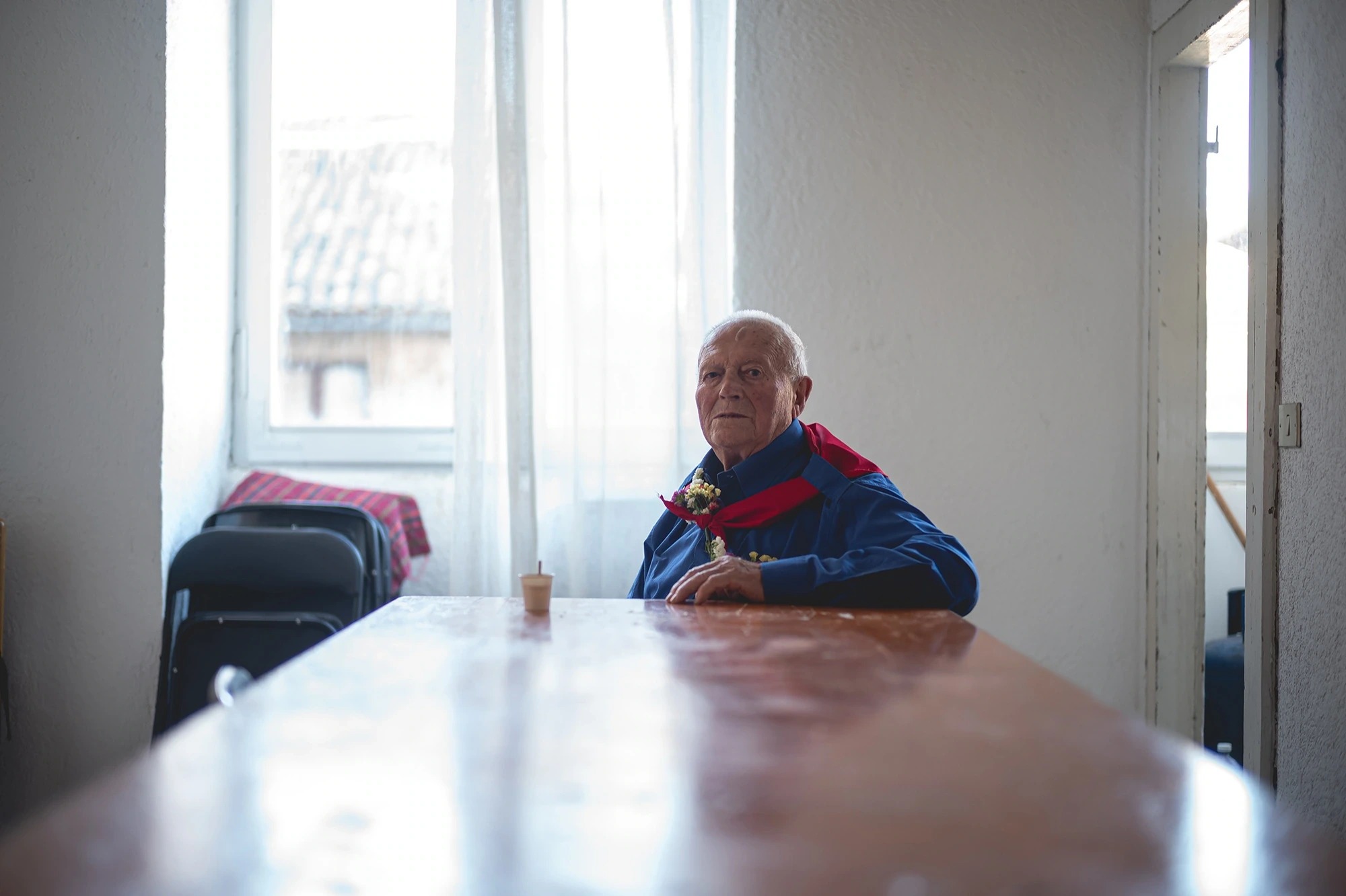 An elderly man with a red sash, alone at a long table by the window.