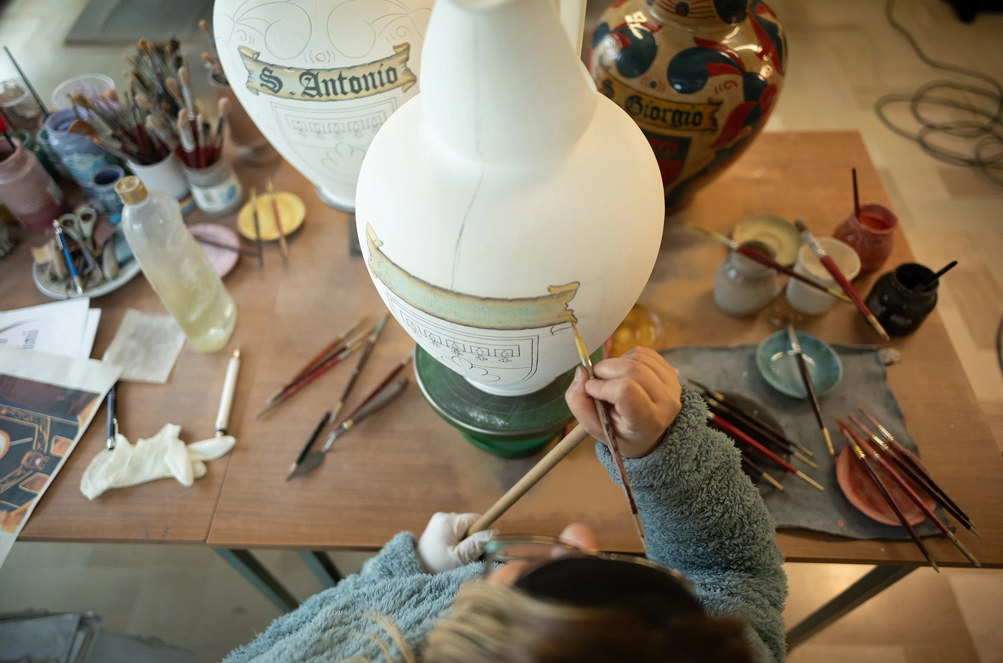 In the workshop, a hand decorates a ceramic amphora lettered «S. Antonio».