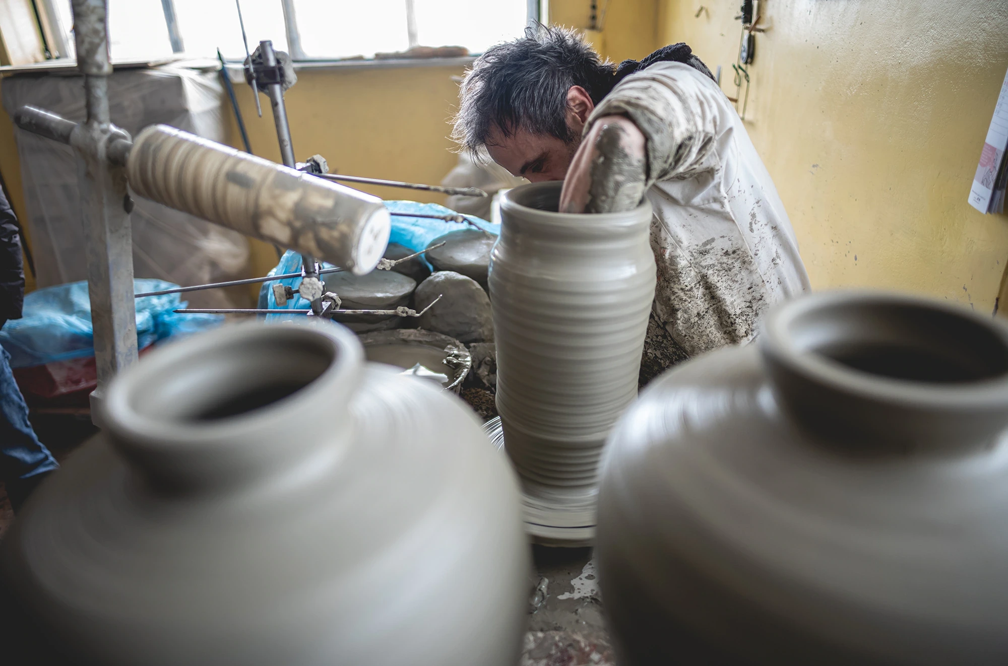 At the wheel, the potter shapes an amphora; others dry alongside.