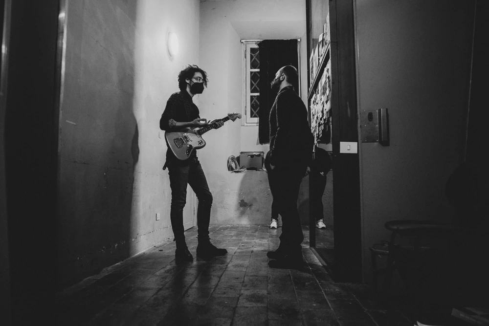 In a corridor, a guitarist plays for someone listening in the doorway.