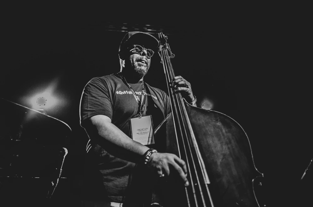 A double bassist in a cap and glasses, lit from the side.