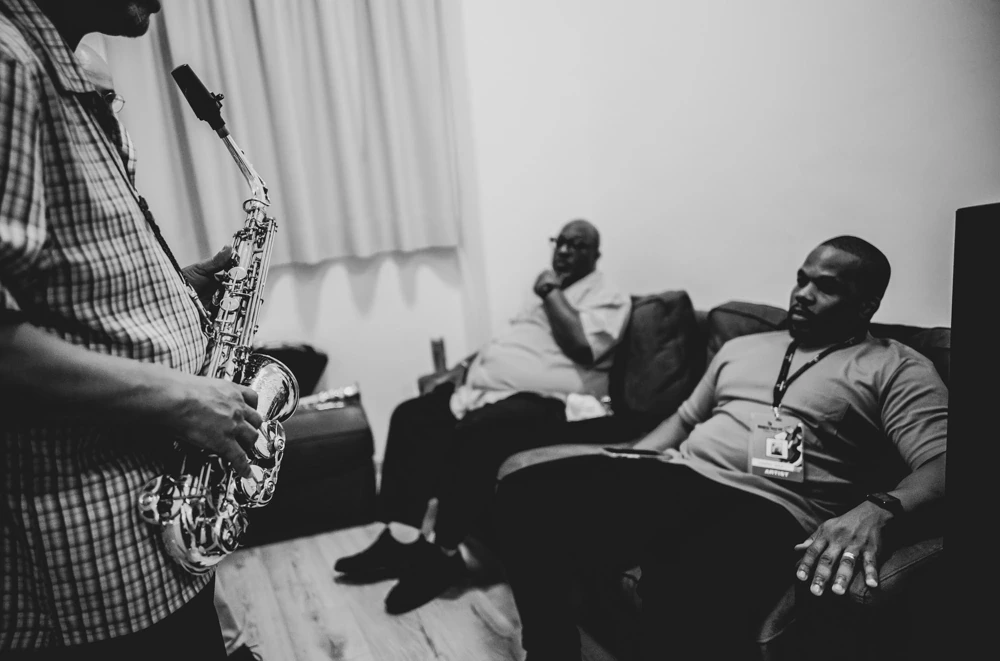 In the dressing room, a saxophonist rehearses while two men listen from the sofa.