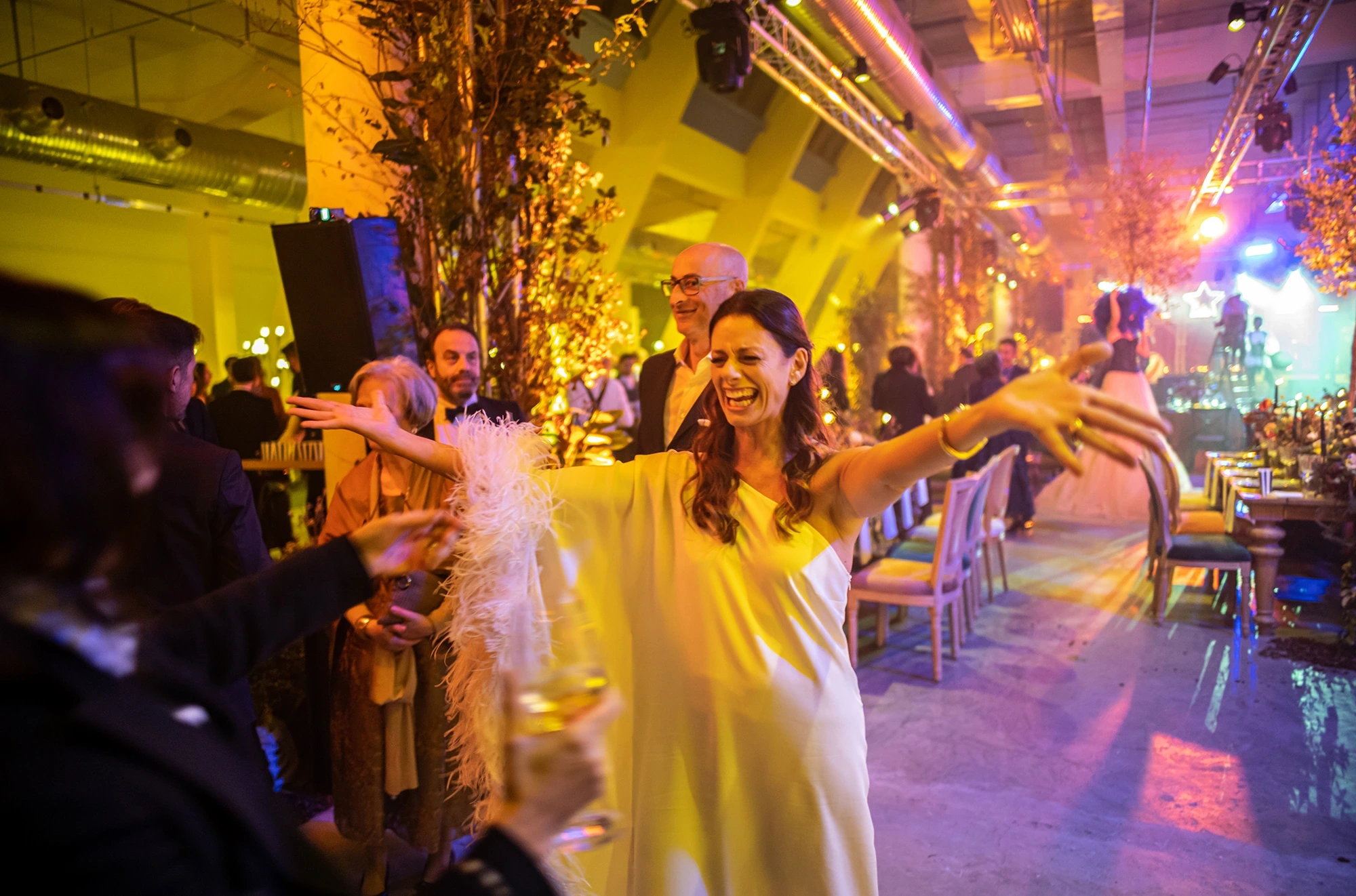 The bride laughs with open arms, in the yellow-lit room.