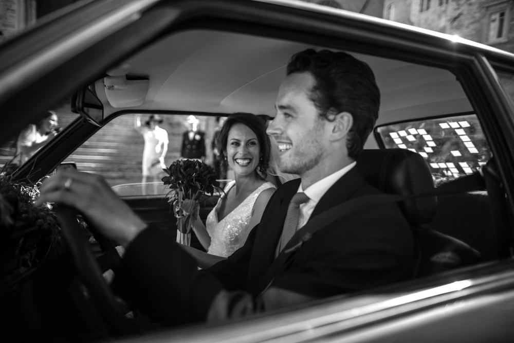 In the car, the bride laughs beside the groom at the wheel.