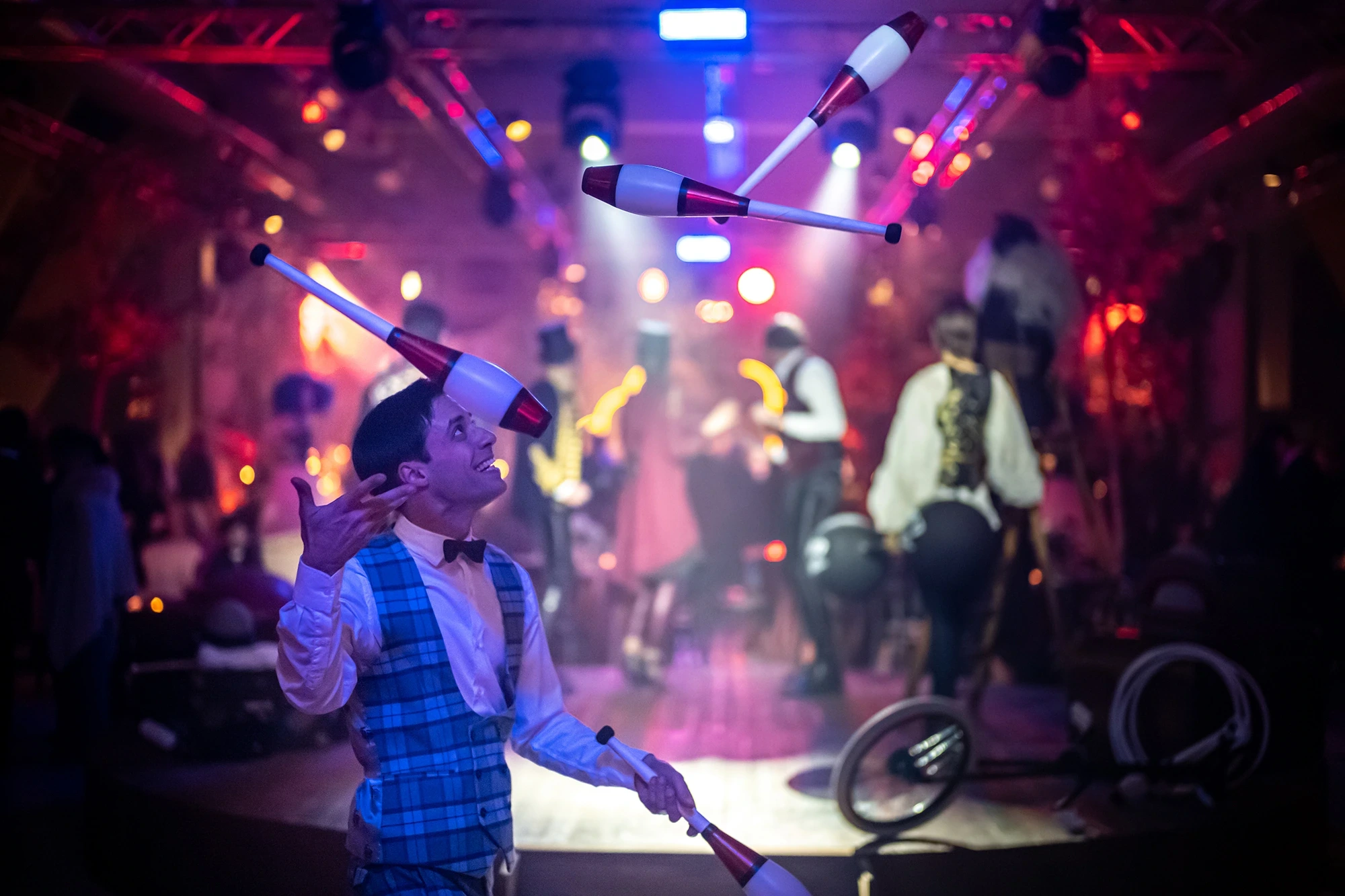 A juggler in a waistcoat throws clubs beneath the lights.